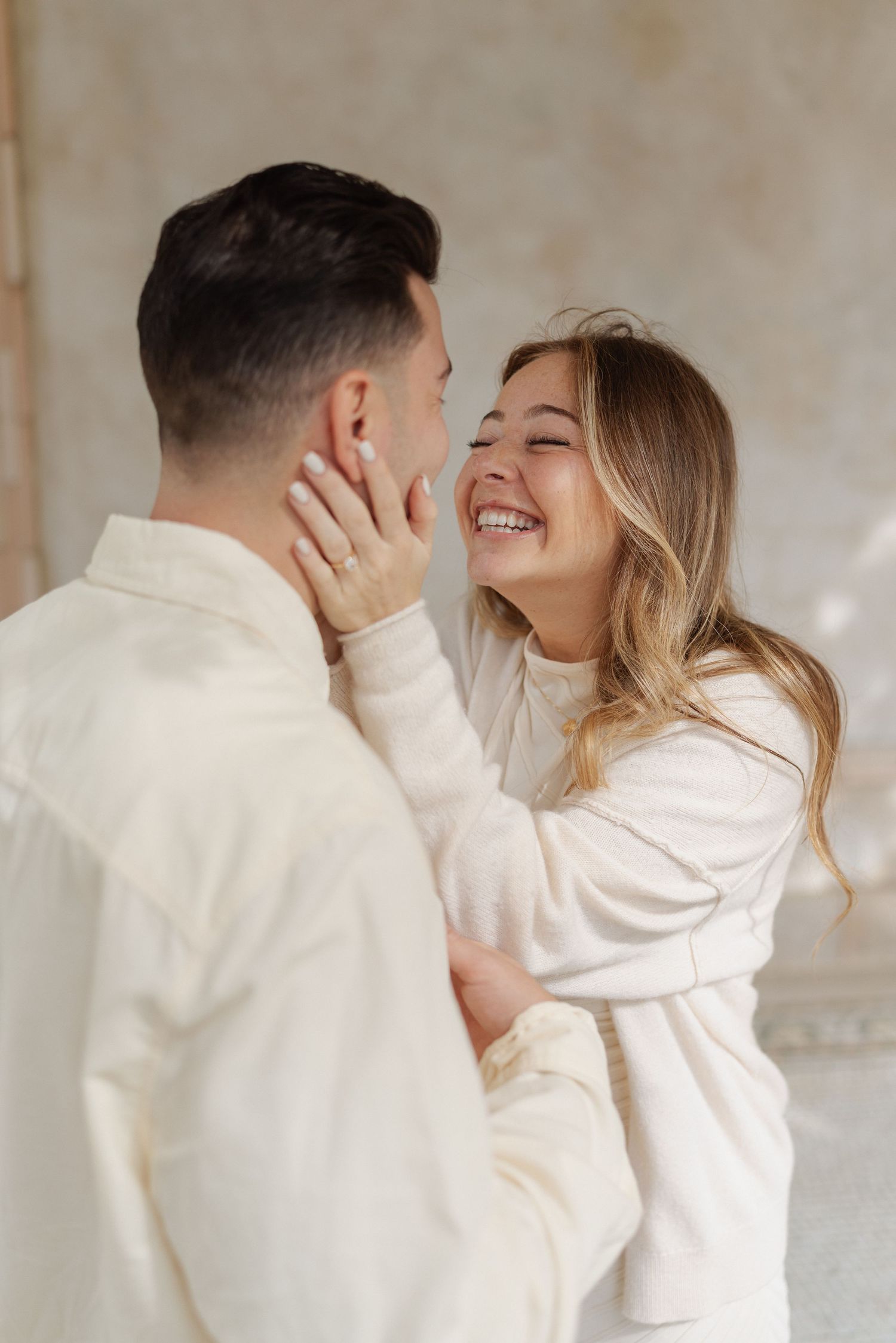 Smiling woman with long blonde hair gently touches a man's face as the couple shares a joyful moment in matching cream outfits.