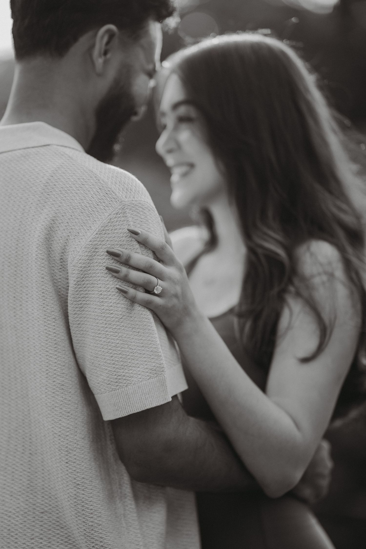 A smiling woman wearing an engagement ring rests her hand on her partners shoulder in black and white.