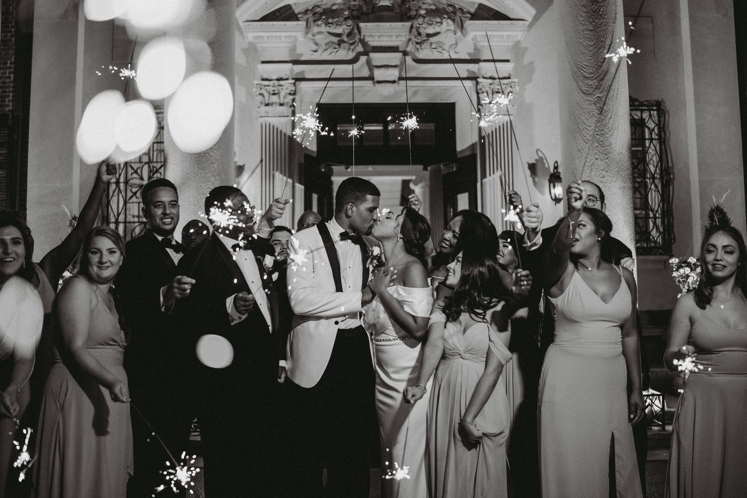 Bride and groom share romantic kiss during sparkler send-off surrounded by cheering wedding guests.