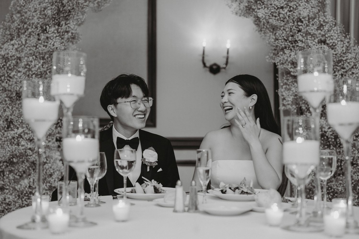 Bride and groom share a joyful moment at candlelit sweetheart table in black and white wedding photo.