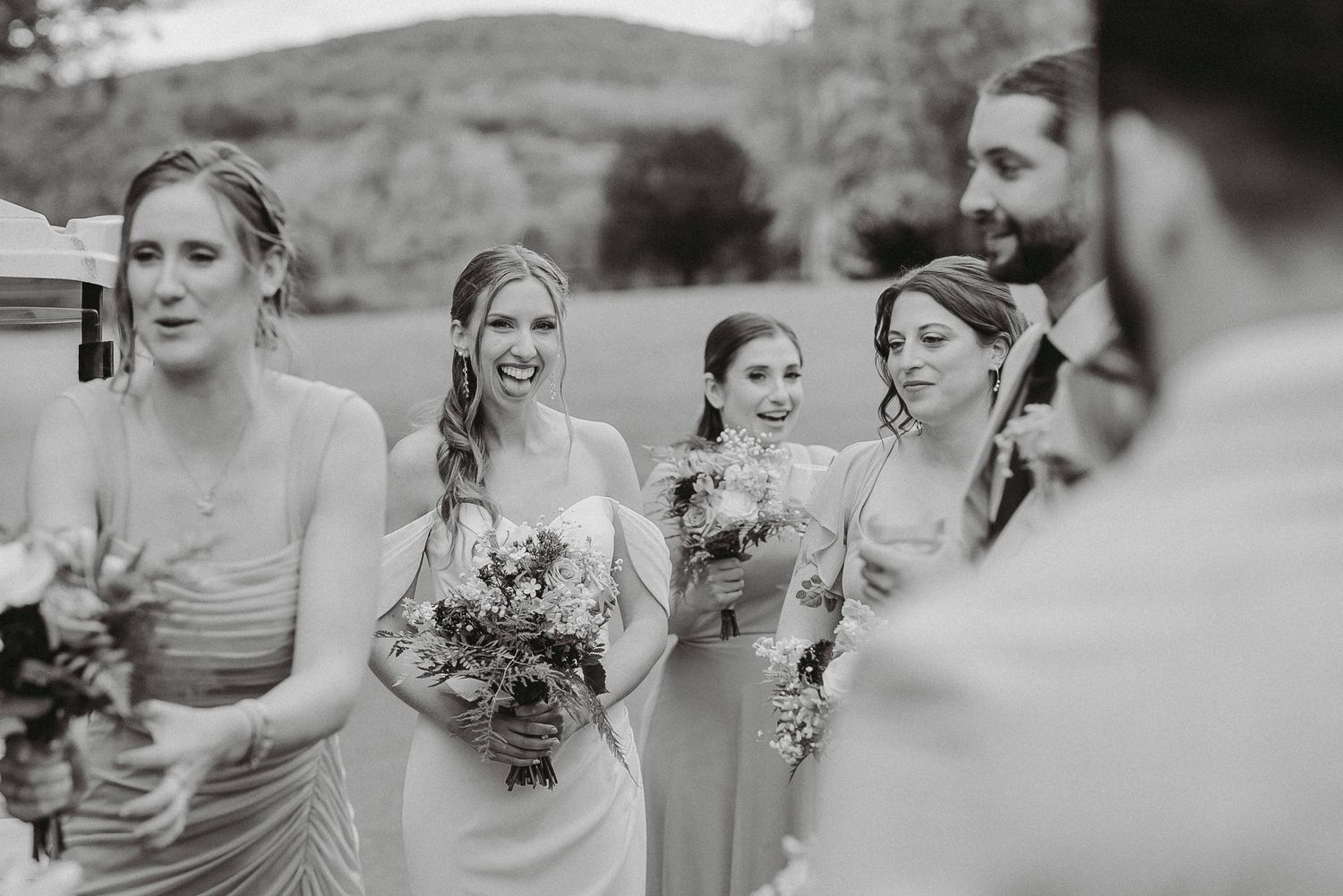 Black and white photo of bridesmaids in gray dresses laughing with bride during outdoor wedding.