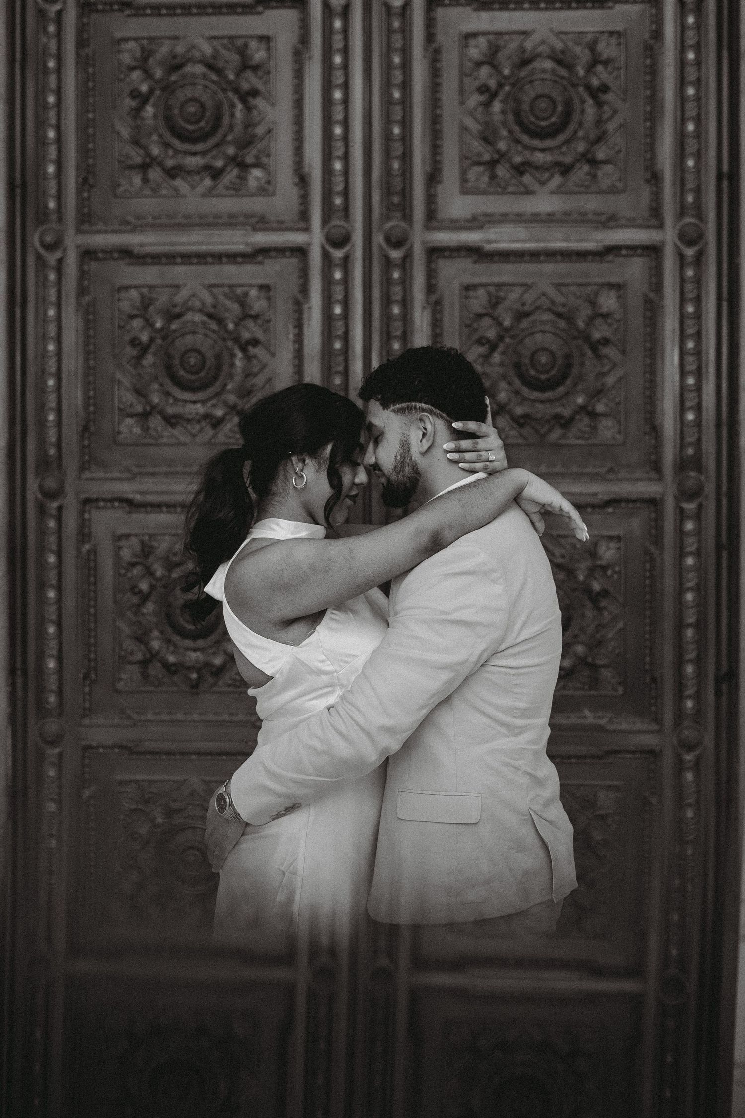 Black and white photo of couple embracing passionately before ornate carved wooden doors.
