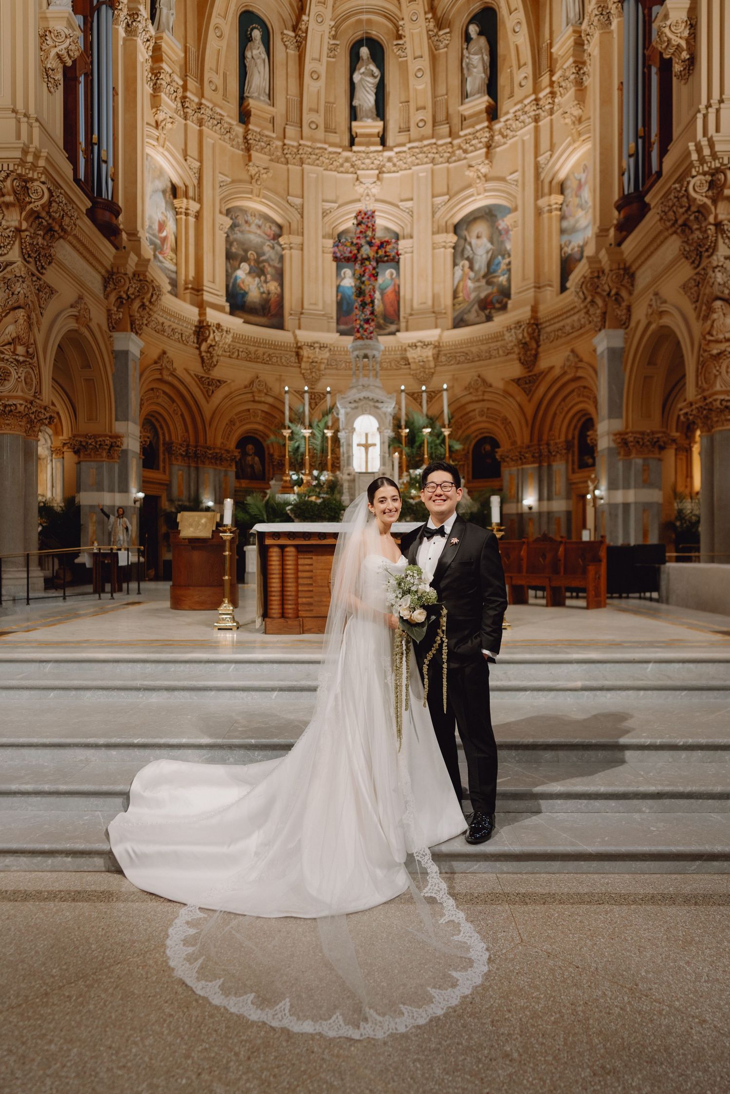 Bride in cathedral veil and groom in tuxedo pose before stunning ornate church altar.