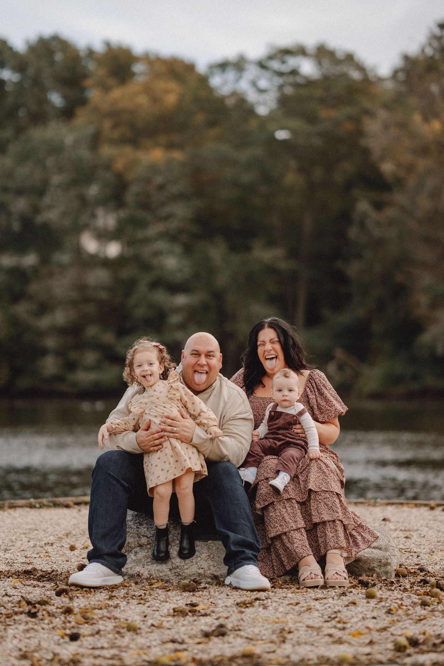 A happy family of four poses and laughs together on a rocky shoreline with autumn trees behind them.