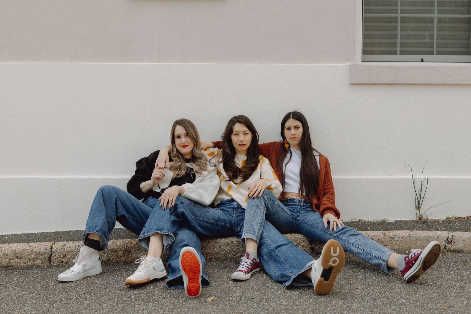 Three young women in jeans sit casually against a white wall outdoors, wearing sneakers and casual tops.
