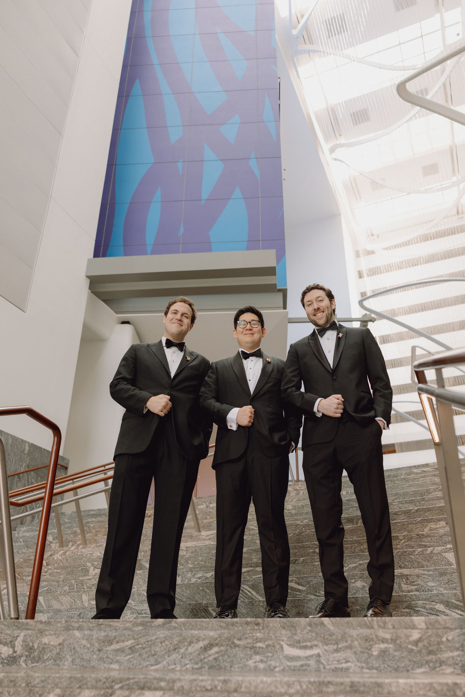 Groom and two groomsmen in black tuxedos pose confidently on marble stairs inside a modern building.