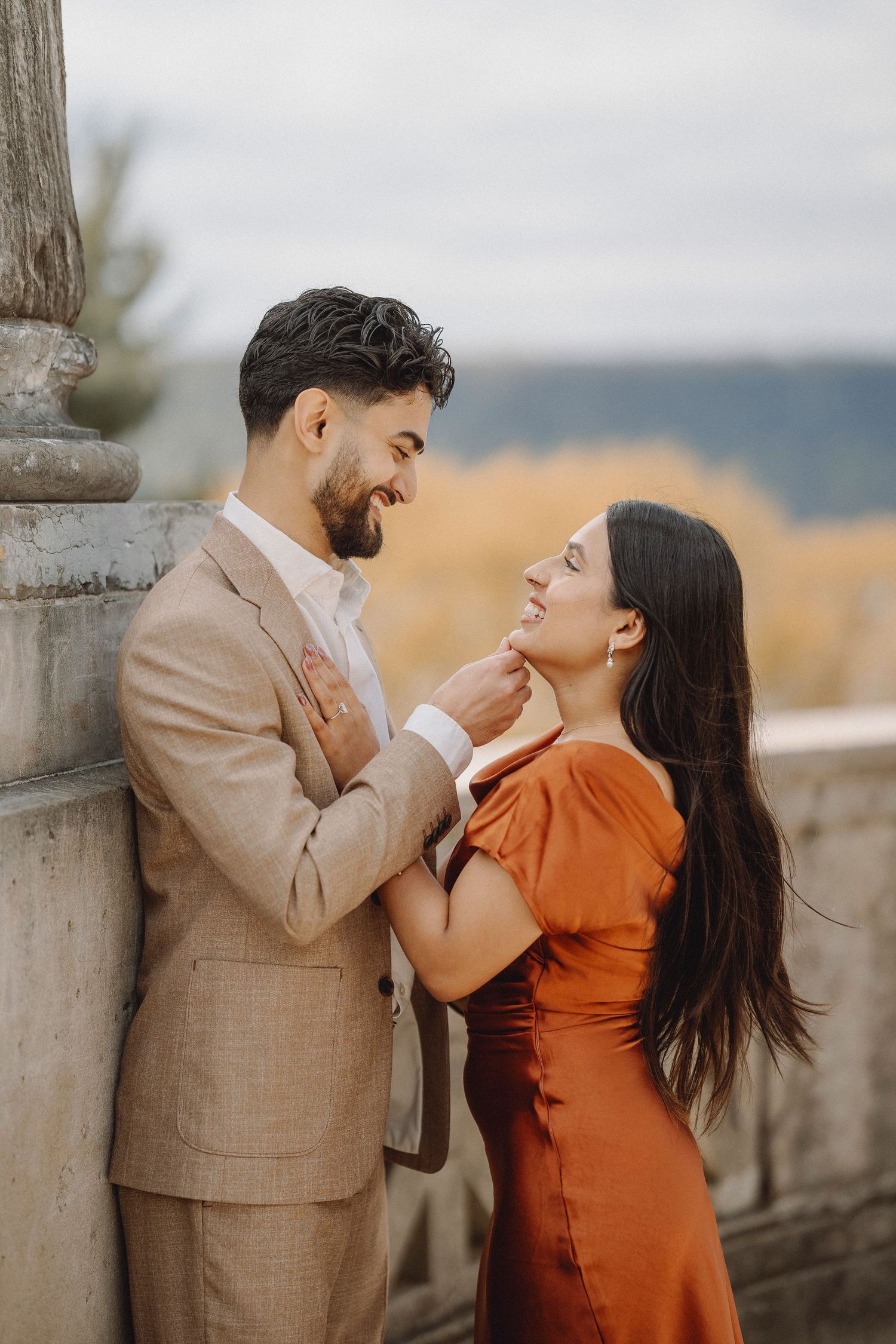 Man in beige suit tenderly touches womans face as they smile together against a stone column.