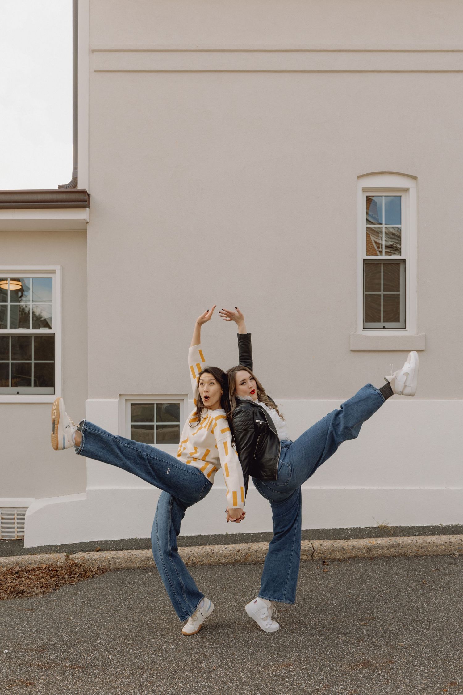 Two young women doing synchronized high kicks in wide-leg jeans outside a beige building.