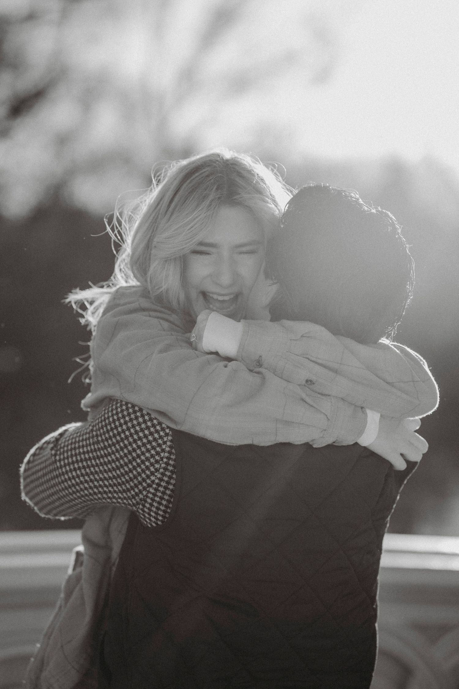 Joyful woman with blonde hair hugging her partner in a sunlit black and white engagement photo.