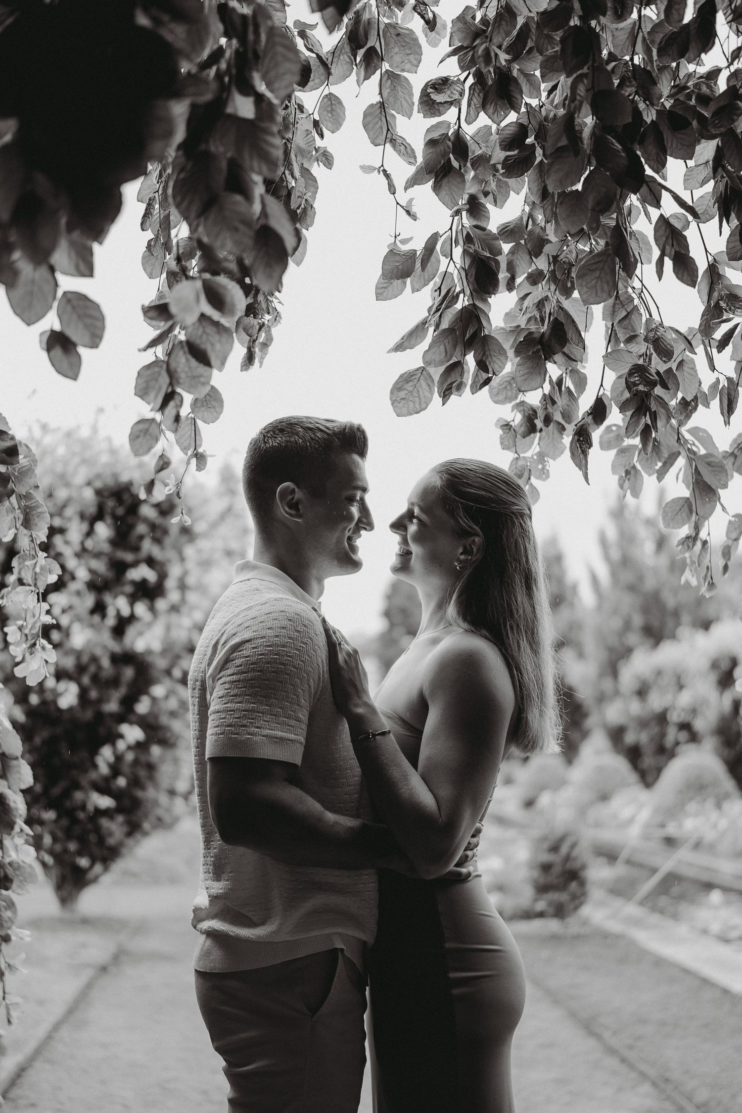 A romantic couple embraces under hanging leaves in a lush garden in black and white.