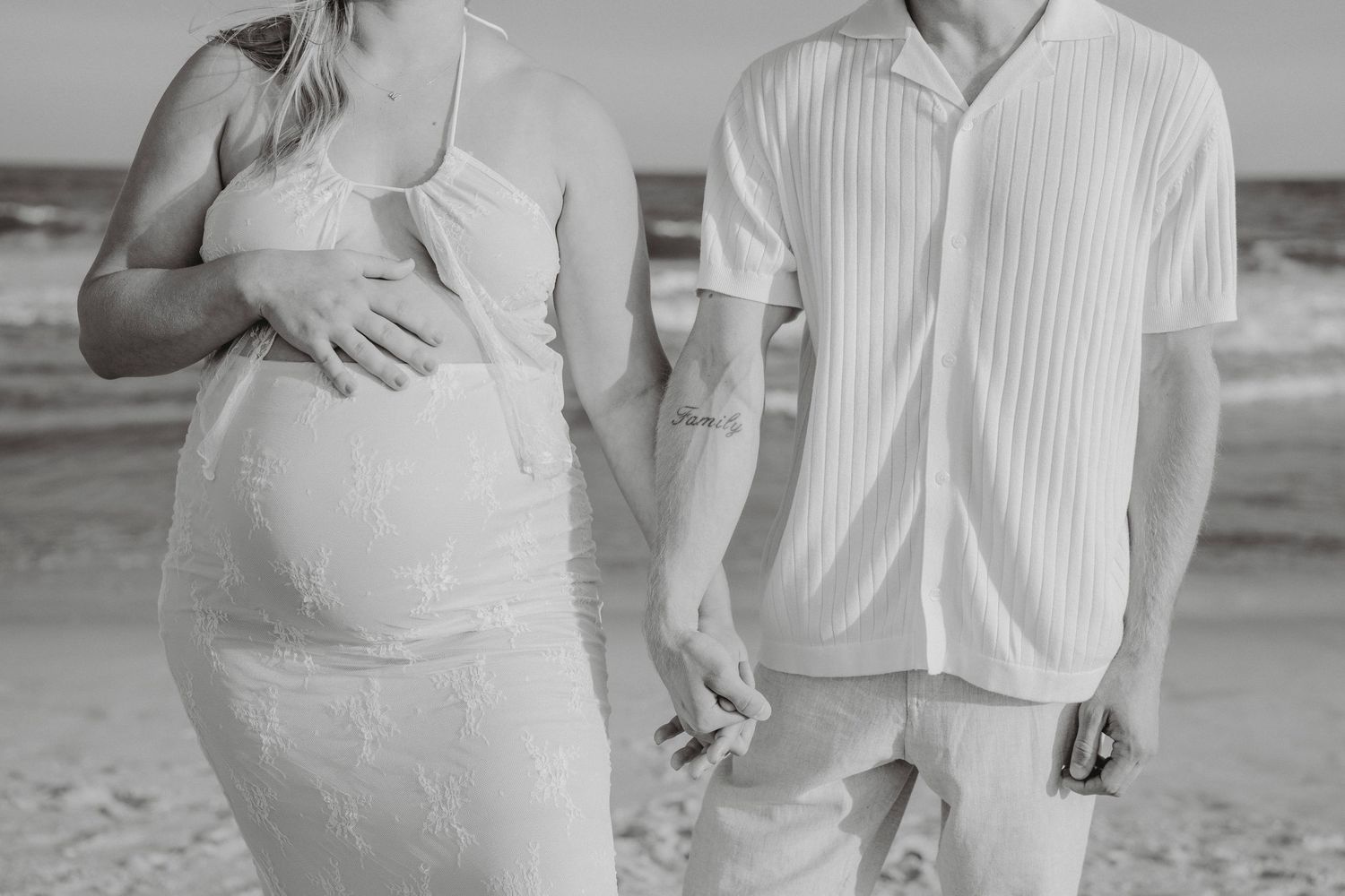 Black and white beach maternity photo of couple holding hands, woman in lace dress cradling baby bump.
