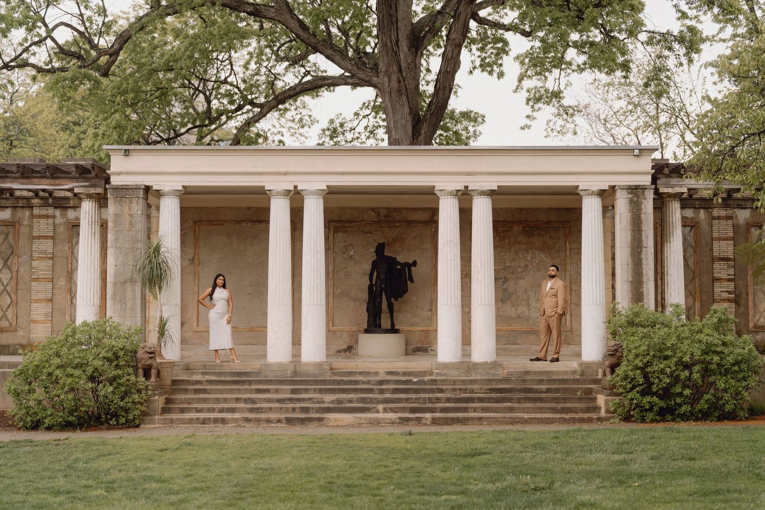 Engaged couple standing apart beneath a classical columned pavilion with a bronze statue centerpiece.