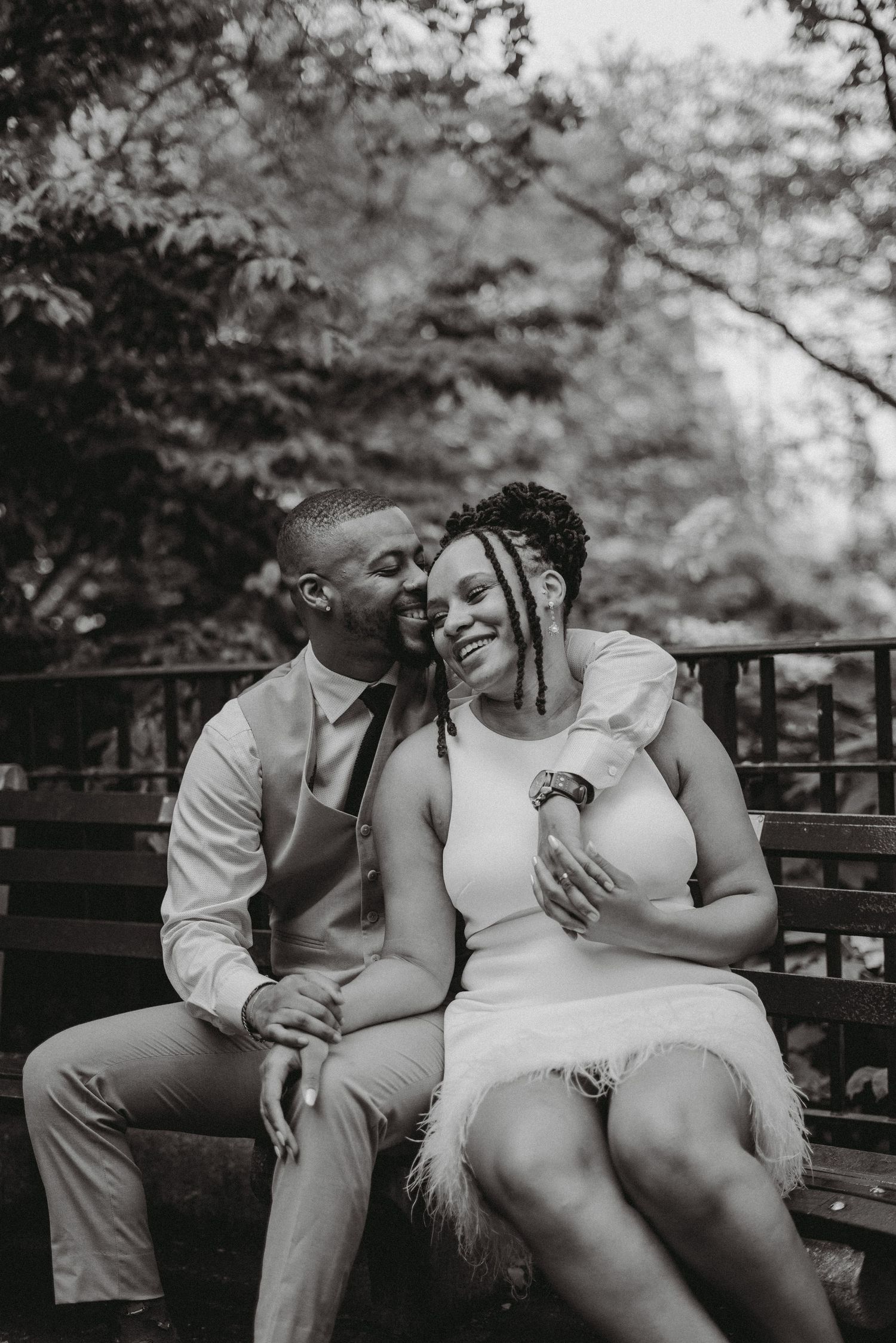 Black and white romantic photo of a couple sitting on a bench outdoors, sharing a tender embrace and smiling.