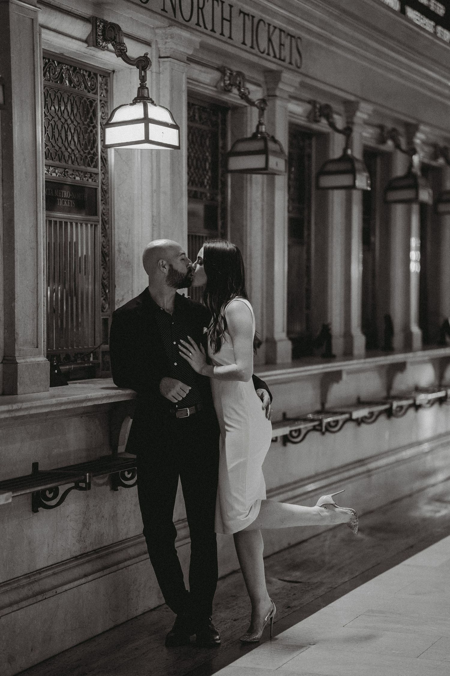 Couple sharing a kiss beside a lit ticket window inside Grand Central Terminal in NYC.
