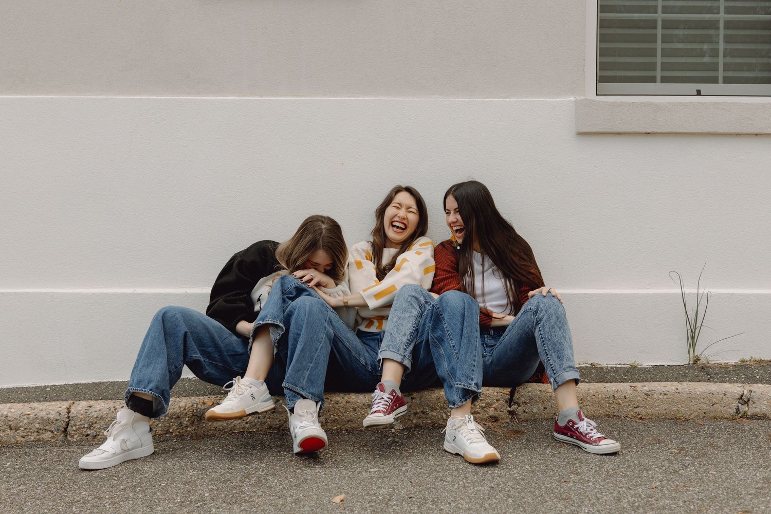 Three young women sitting on a curb laughing together, wearing jeans and sneakers against a white wall.