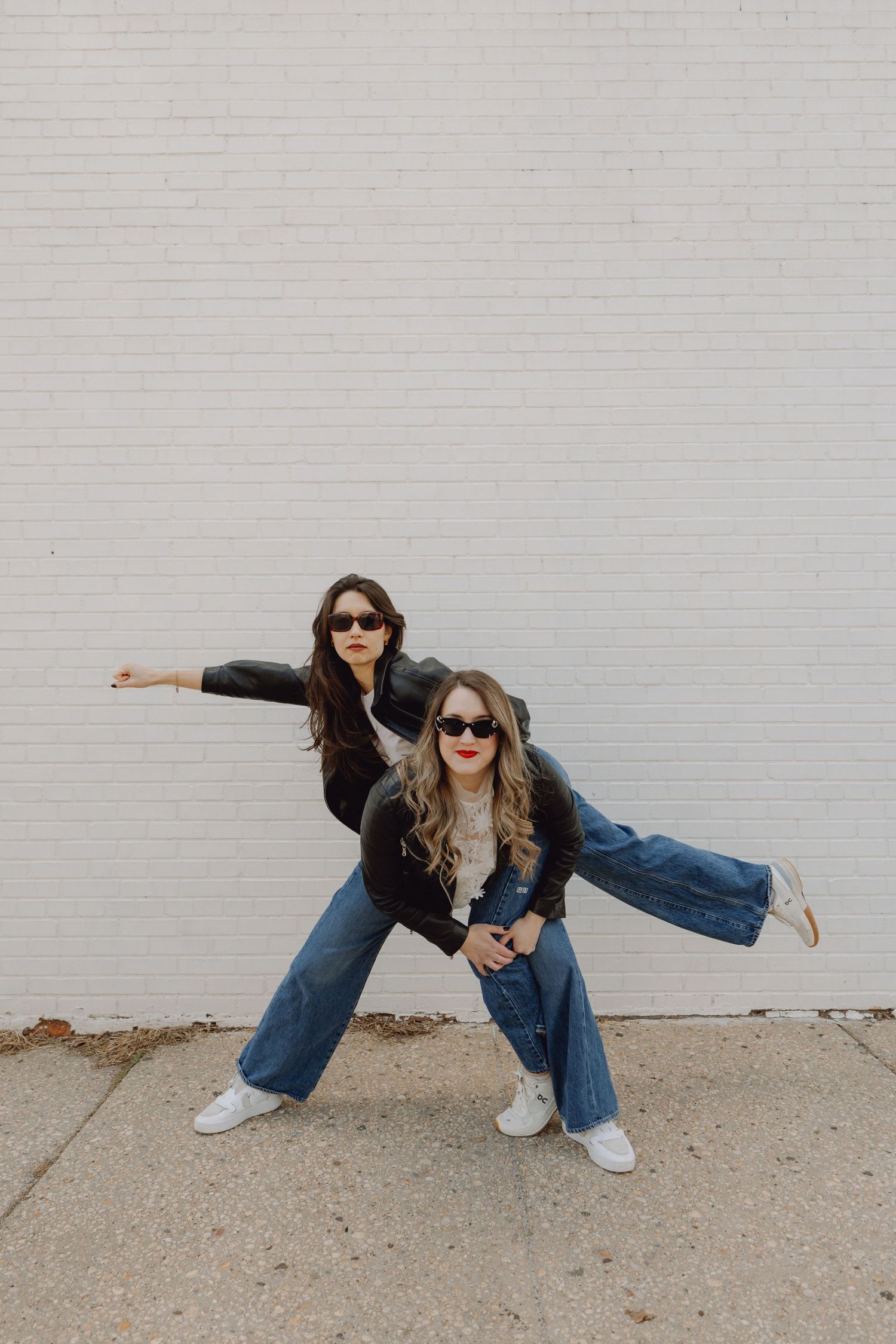 Two women in matching black leather jackets and wide-leg jeans posing dynamically in front of a white brick wall.