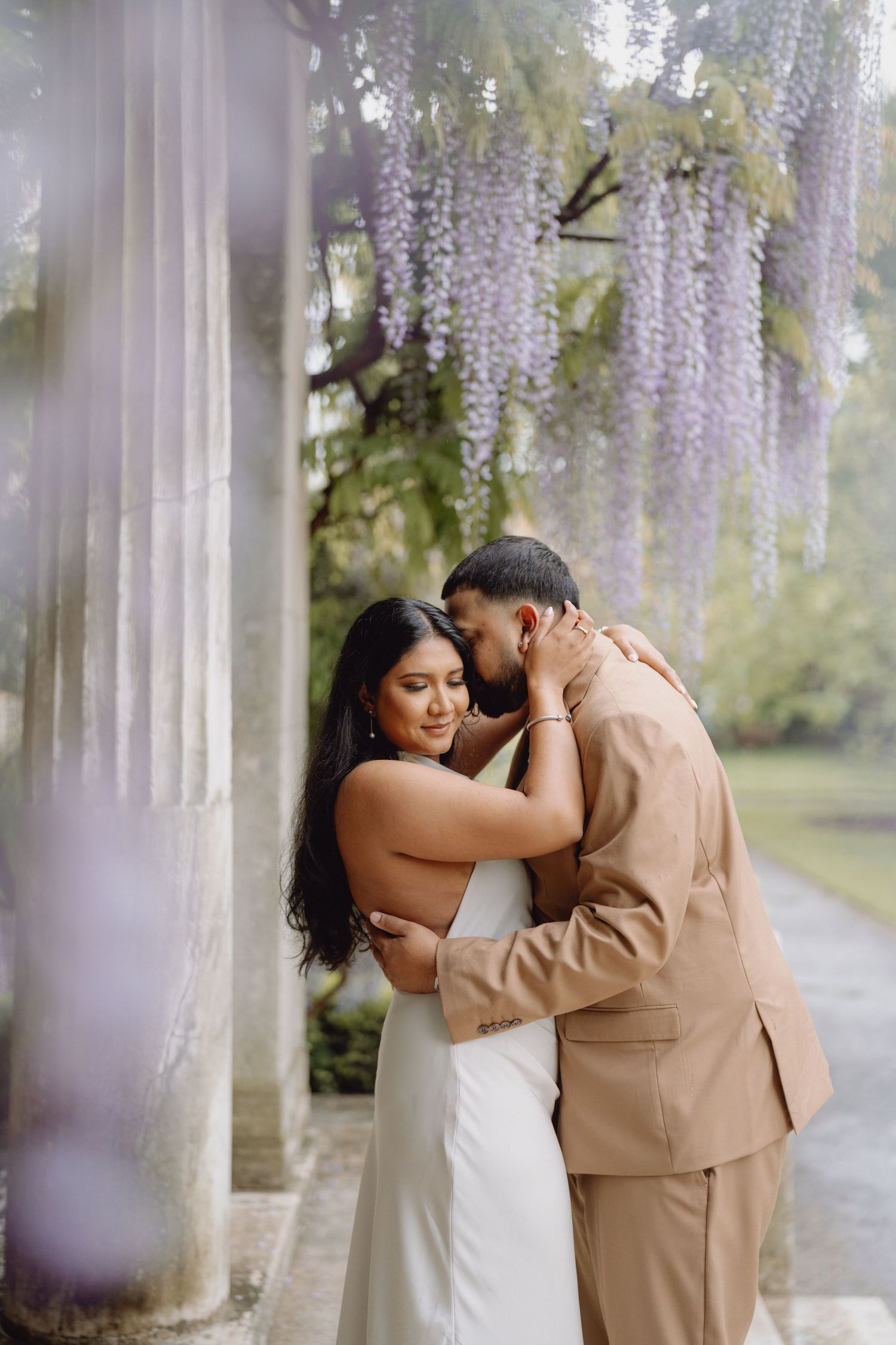 Couple embracing romantically under hanging purple wisteria blooms between stone garden columns.