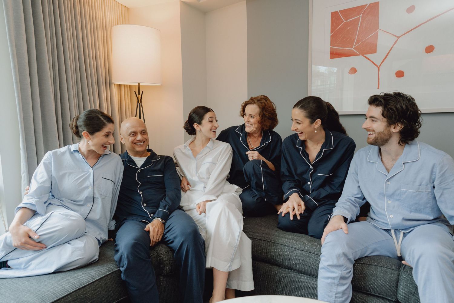 Bride and wedding party laugh together in matching pajamas while seated on a hotel room couch.