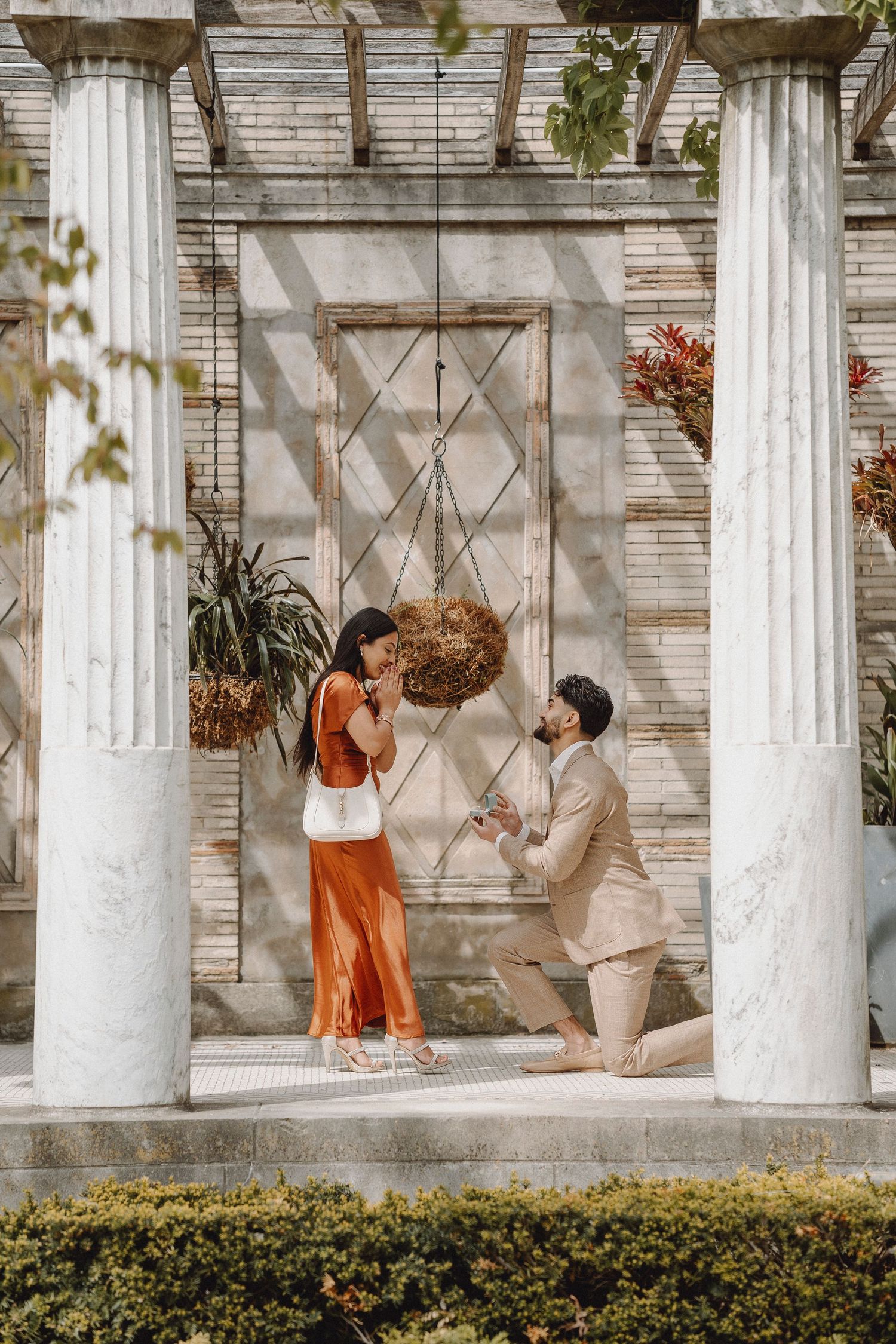 Man proposes on one knee to surprised woman in orange dress under a garden pergola with columns.