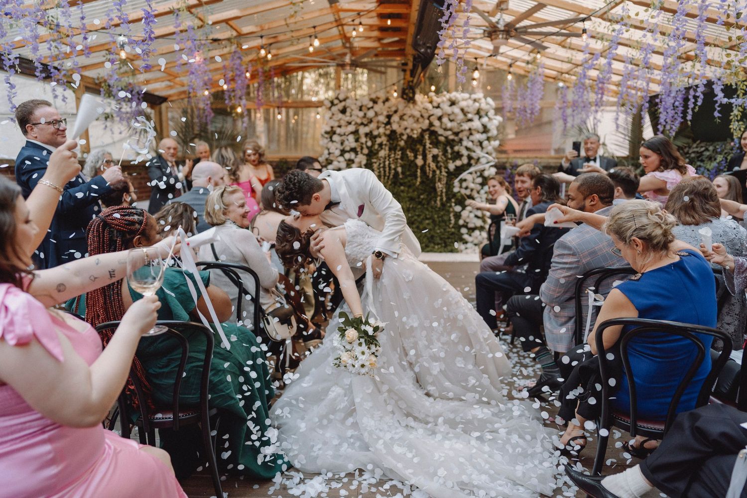 Bride and groom share a kiss while confetti falls during their wedding ceremony in a floral greenhouse venue.