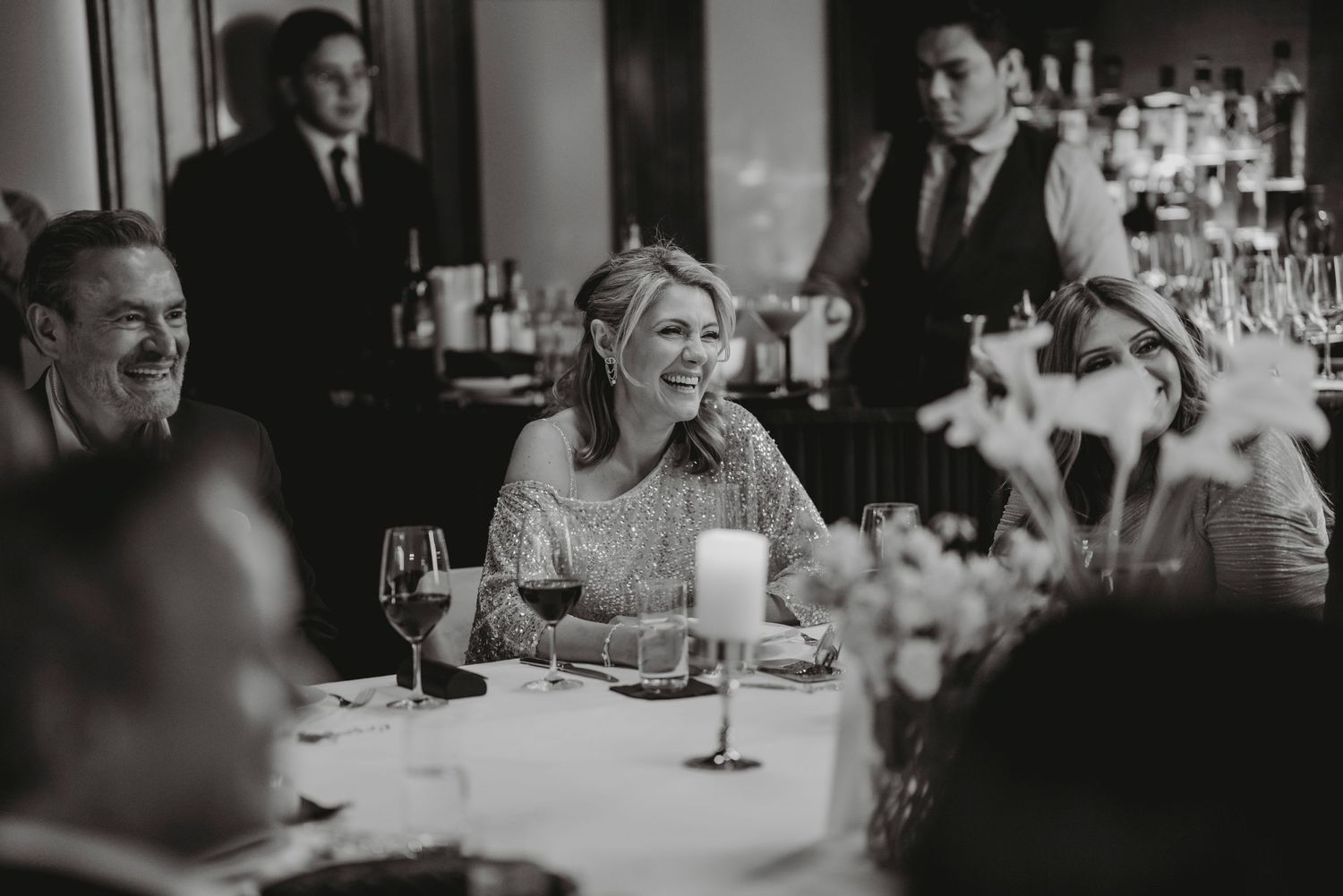 Black and white photo of a smiling woman in sequined dress enjoying a wedding reception dinner with guests.