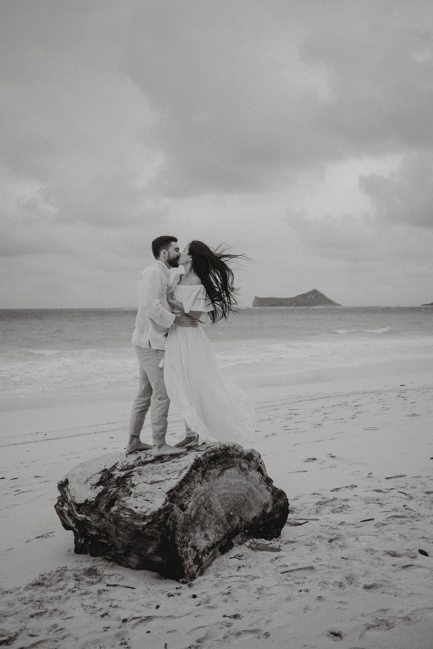 Couple kissing on driftwood log on dramatic beach with island visible in black and white.