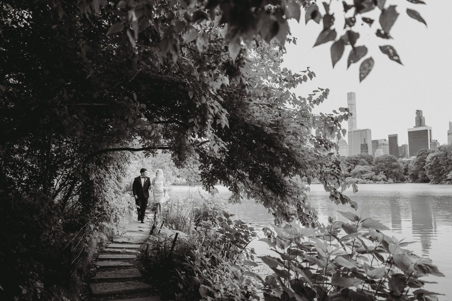 Bride and groom walk along a scenic Central Park lakeside path with NYC skyline in background, black and white.