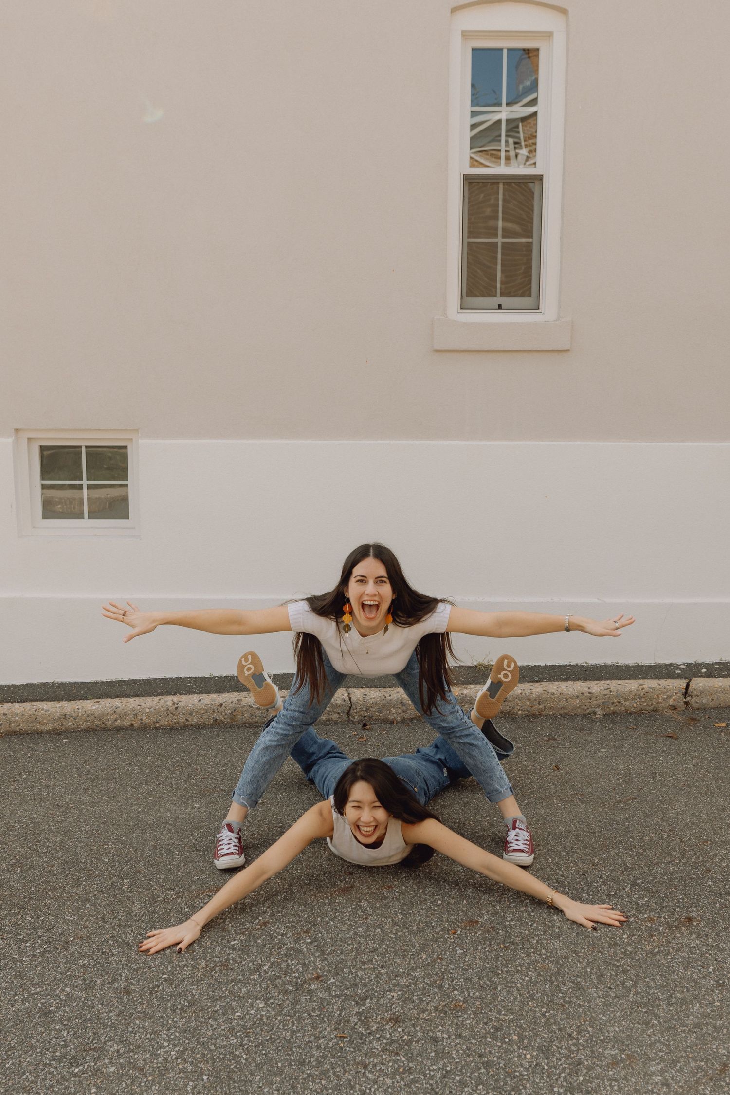 Two young women posing playfully outdoors, one lying on ground with arms spread, other sitting on her back with arms outstretched.