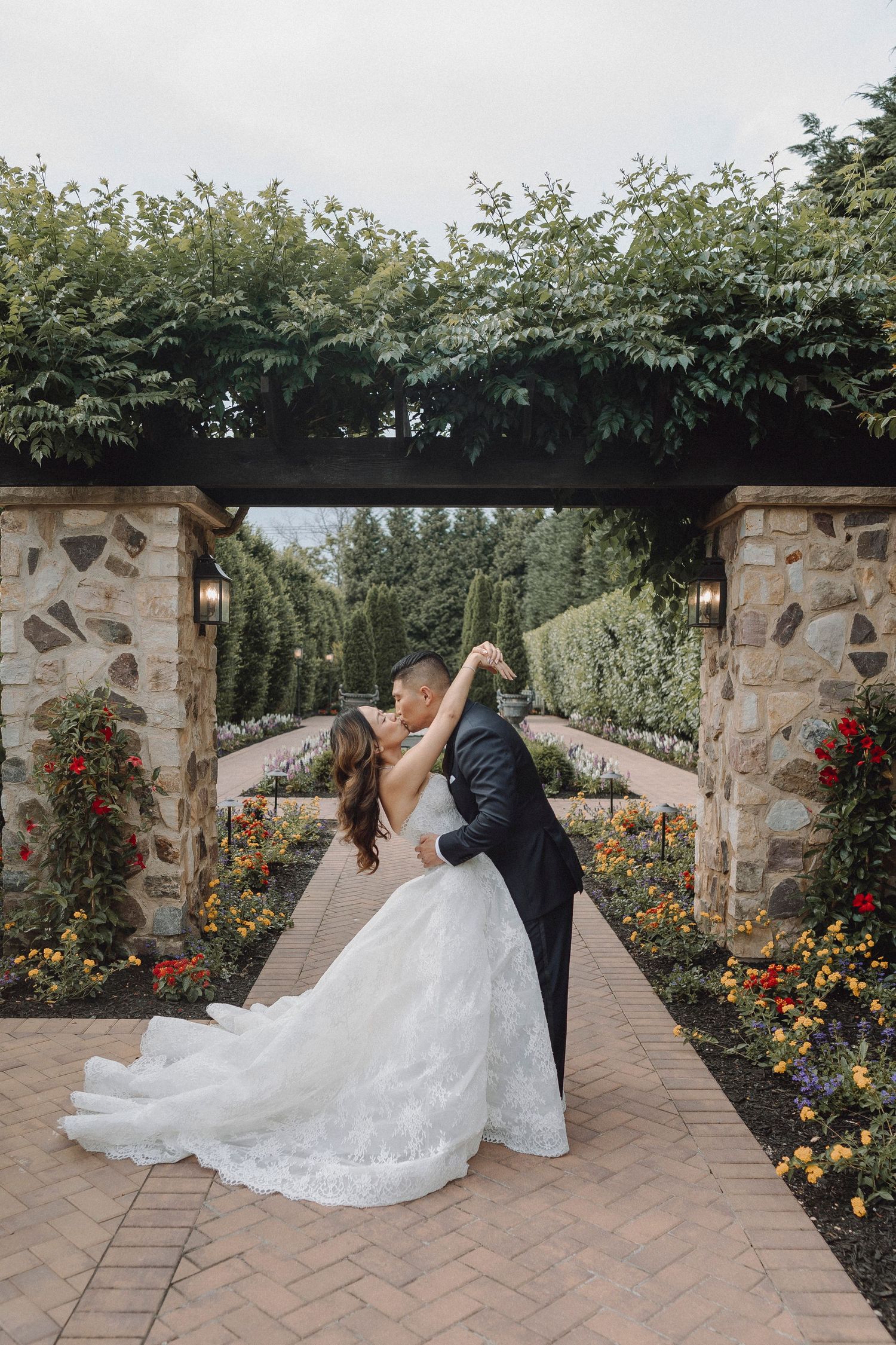 Bride and groom share a romantic dance under a stone pergola surrounded by colorful garden flowers.