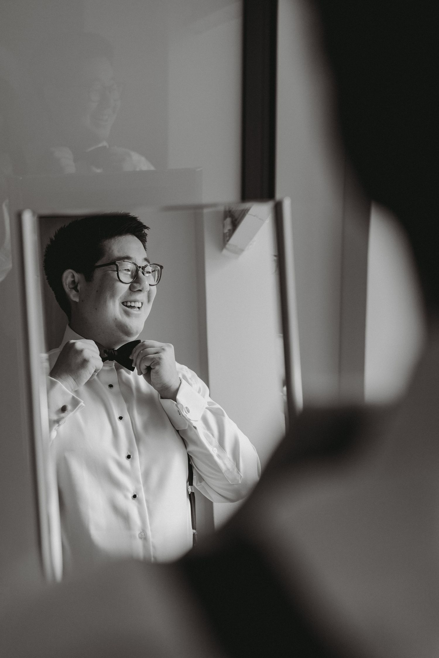 Groom smiling in mirror adjusting bow tie in black and white wedding preparation photo.