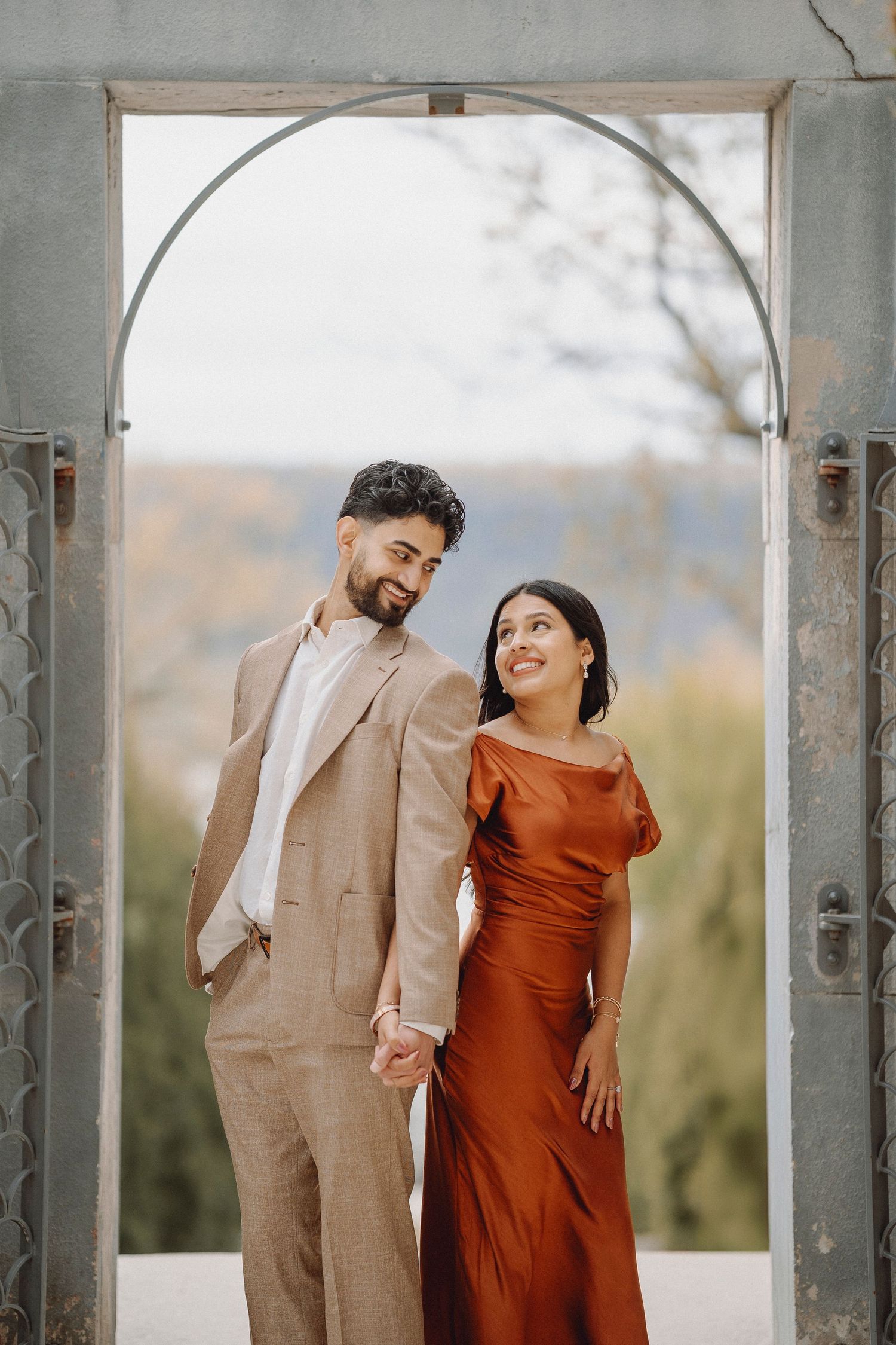 Smiling couple holding hands framed by a stone archway with autumn foliage in the background.