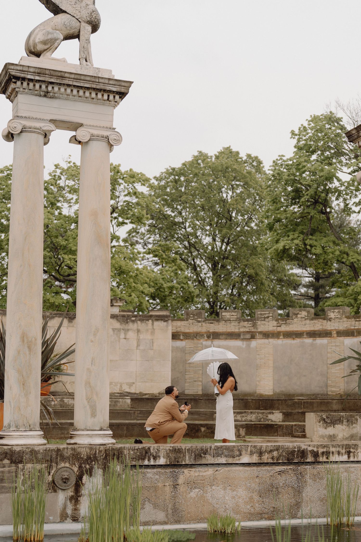 Man kneels in a marriage proposal before a woman in a white dress holding an umbrella beside stone columns, a pond and trees.