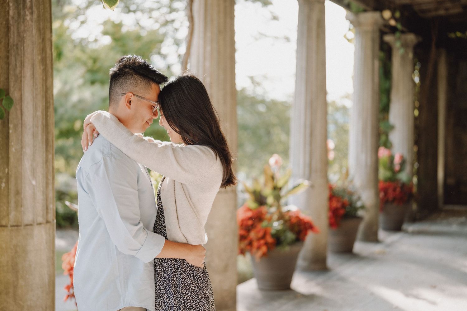 Romantic couple embracing forehead to forehead on a sunlit stone patio lined with columns and vibrant red flower pots.