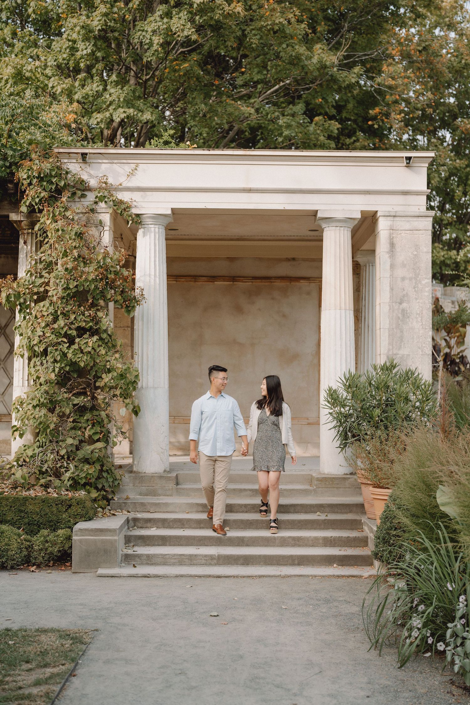 Happy couple holding hands while walking down steps of an elegant garden pavilion with white columns.