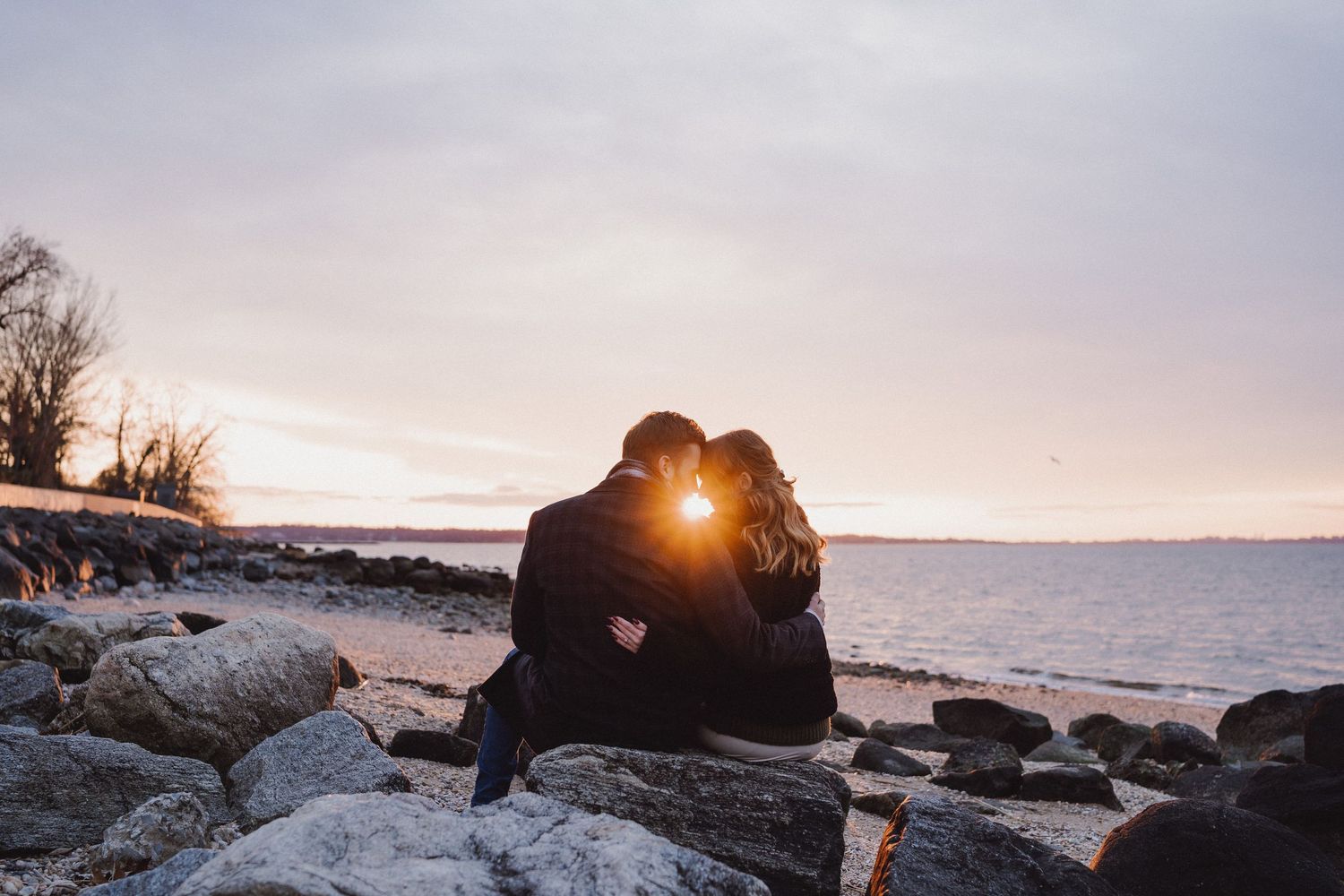 Couple sitting on rocks sharing a kiss at sunset by the water.
