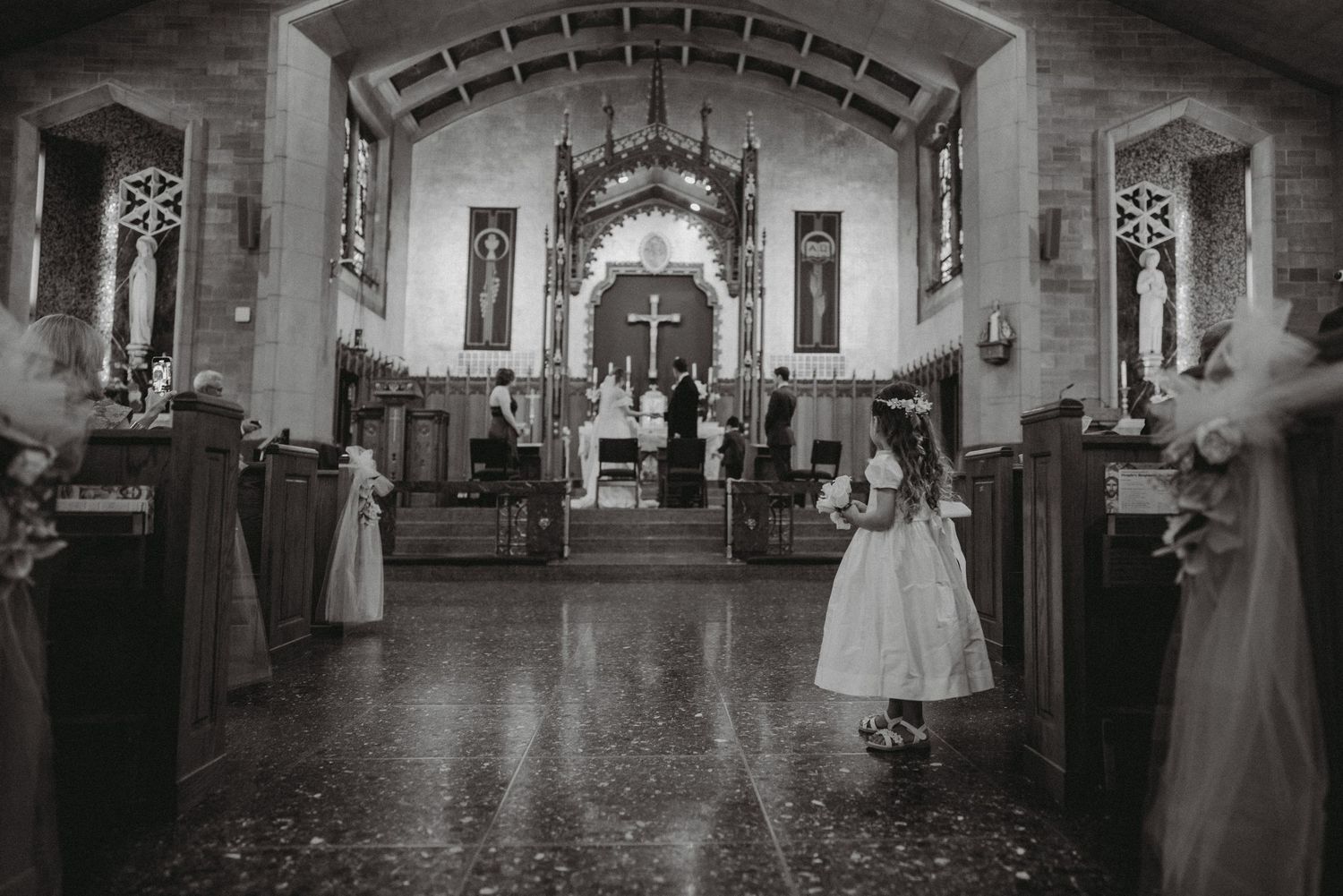 Flower girl stands in church aisle during wedding ceremony with bridal party at the altar.