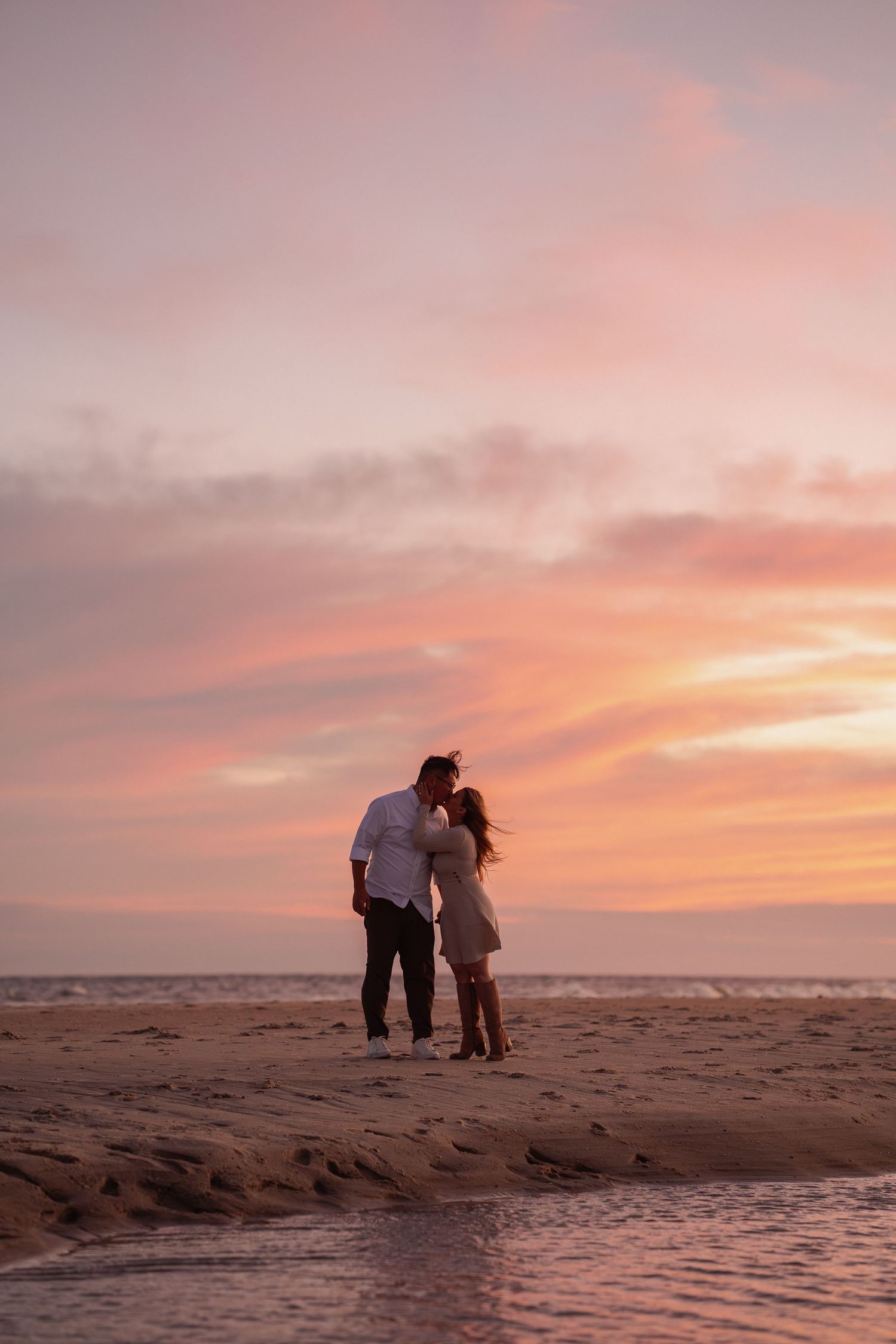 Couple sharing a romantic kiss on a sandy beach during a stunning pink and orange sunset.