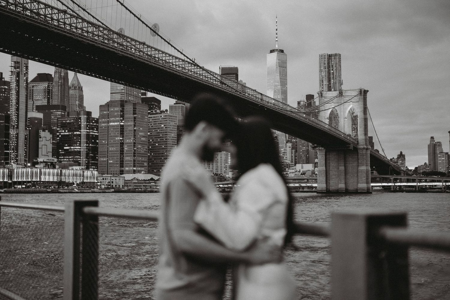 Black and white photo of a couple embracing near the waterfront with the Brooklyn Bridge and Manhattan skyline softly blurred behind them.