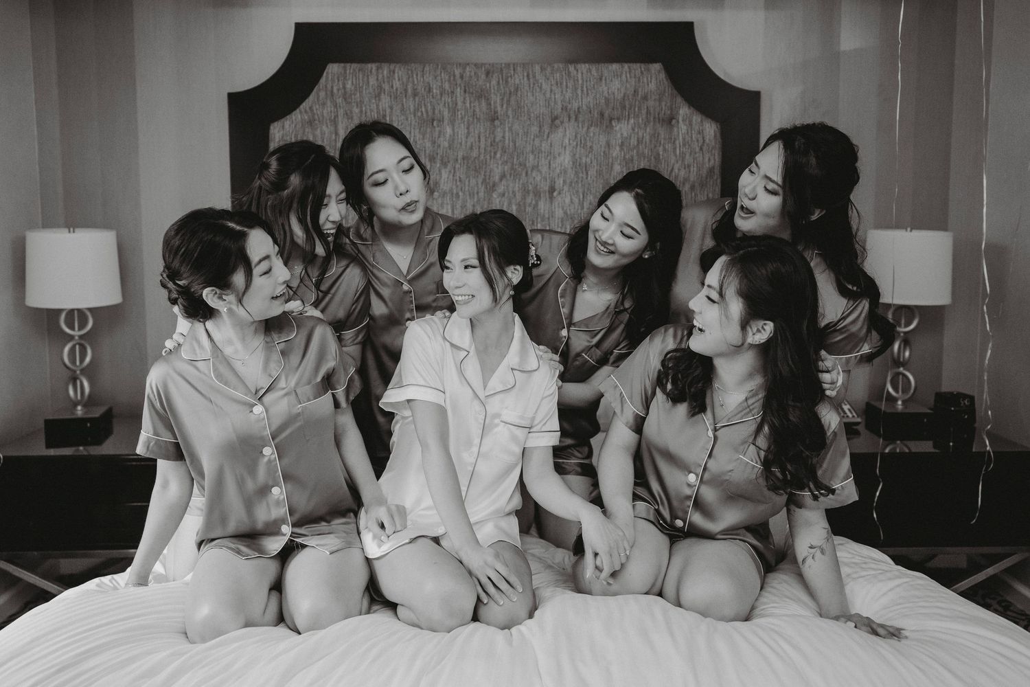 Black and white photo of bride with bridesmaids in satin pajamas on hotel bed.