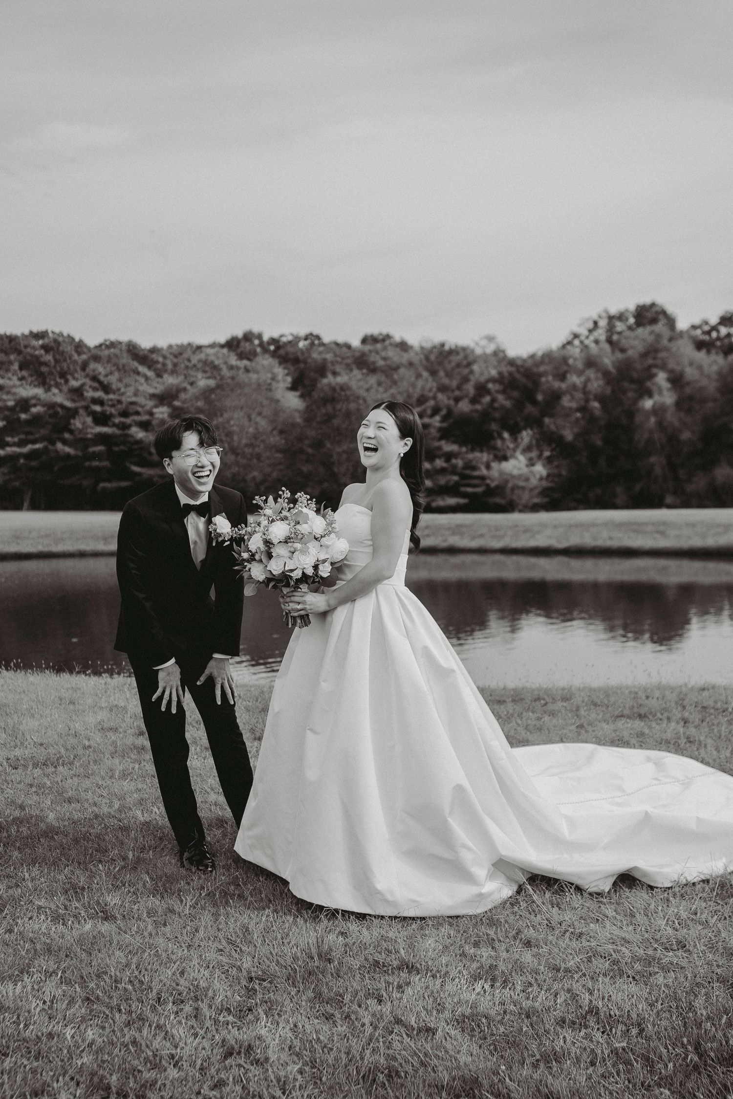 Bride and groom laugh joyfully together outdoors holding bouquet near pond in black and white wedding photo.