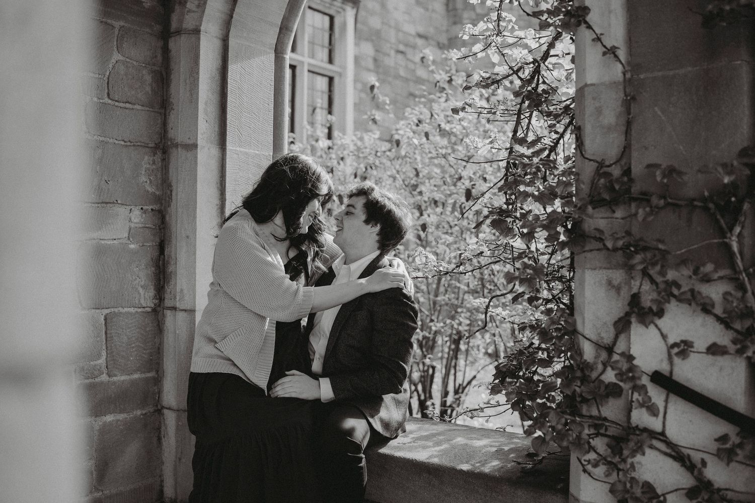 Black and white photo of a couple sharing a romantic moment against a stone building wall.