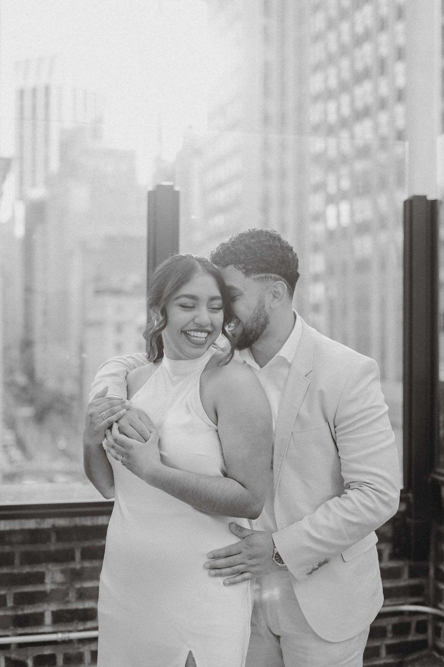 Black and white rooftop photo of a couple embracing, man whispering to laughing woman with city skyline behind them.