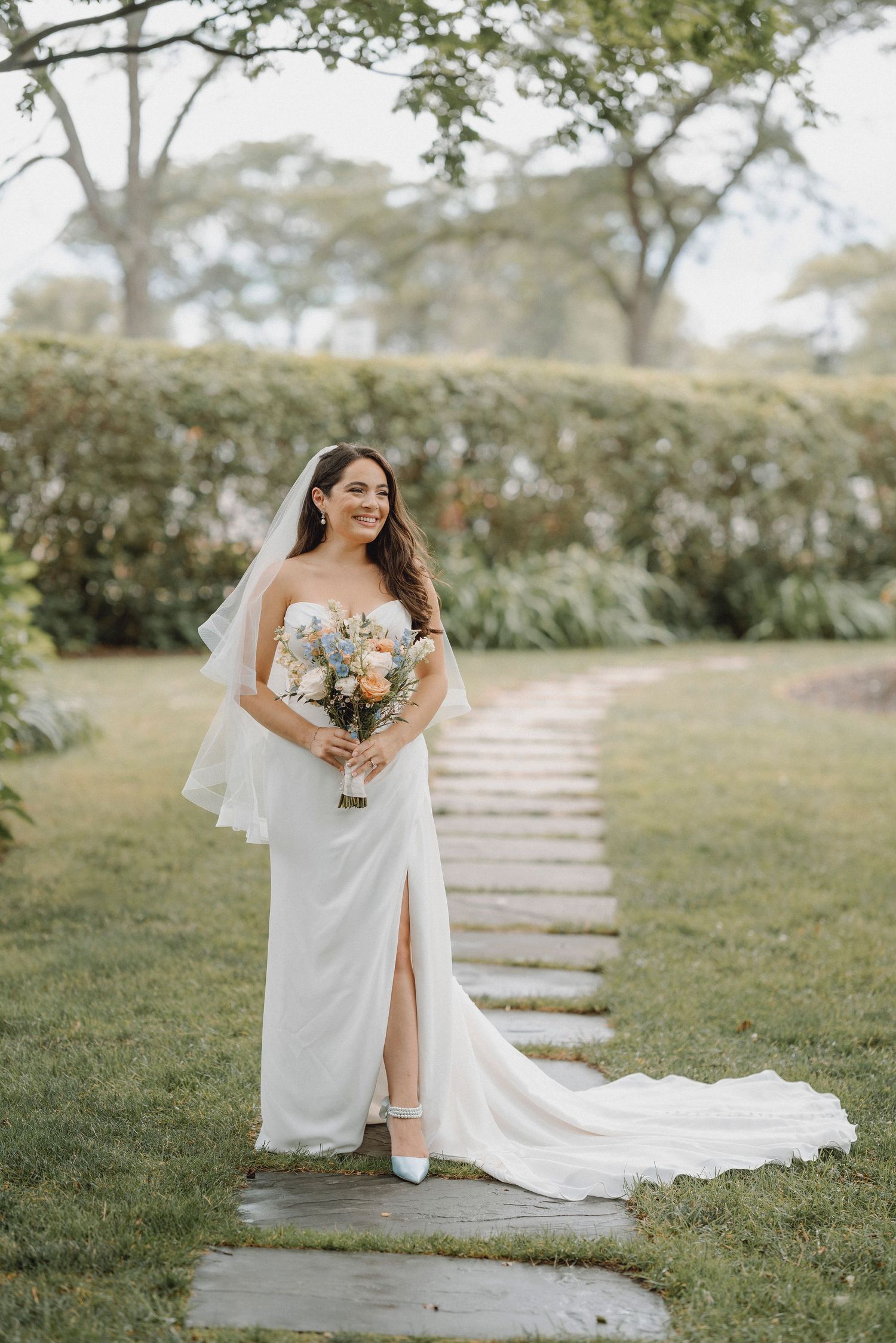 Bride in a white strapless gown with a veil holding a pastel bouquet on a garden stone pathway.