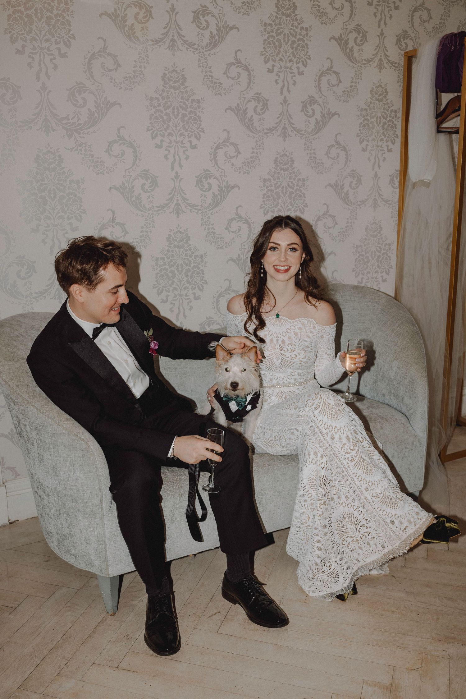 Smiling bride in lace gown and groom in tuxedo seated with their decorated dog and champagne glasses.