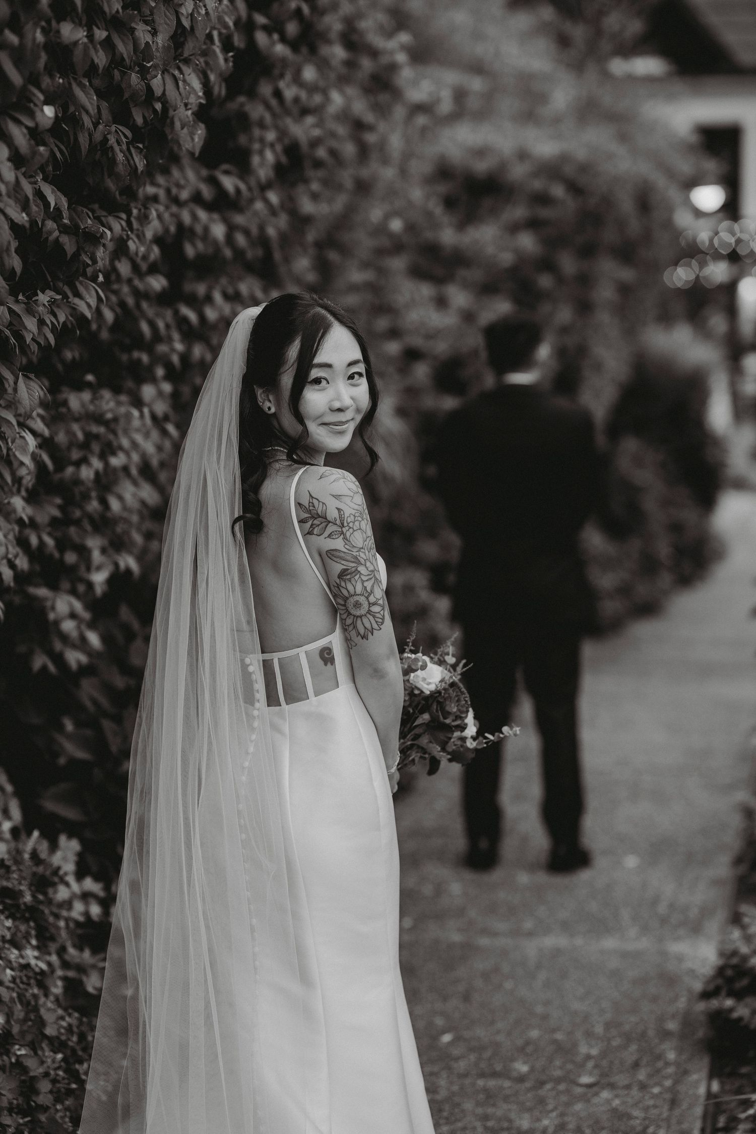 Bride in backless gown with tattoo and veil smiles over shoulder in black and white portrait.