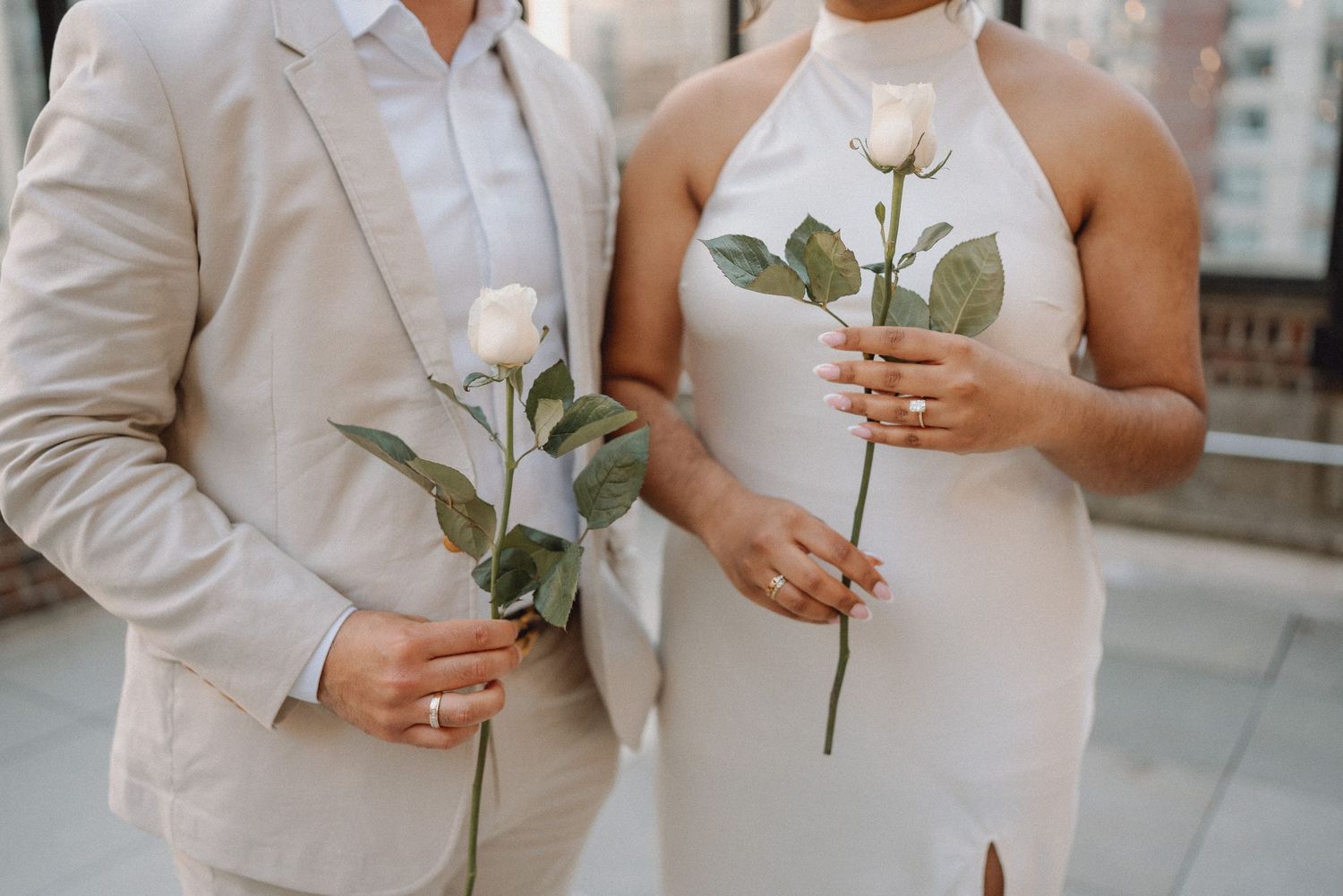Newlyweds holding single white roses in elegant wedding attire on a rooftop venue.