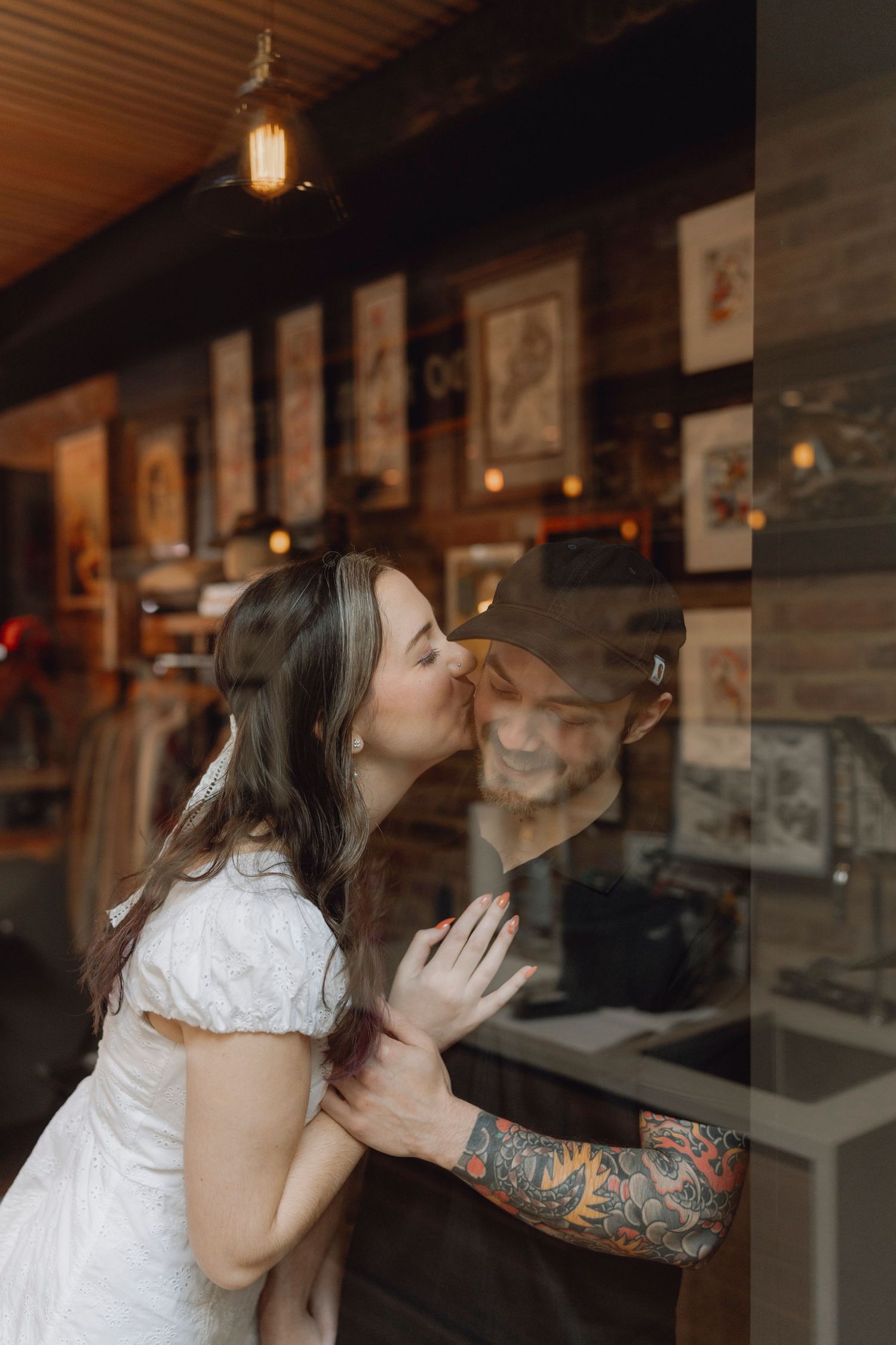 Woman in white dress kissing her tattooed partner through a glass window of a cozy shop.