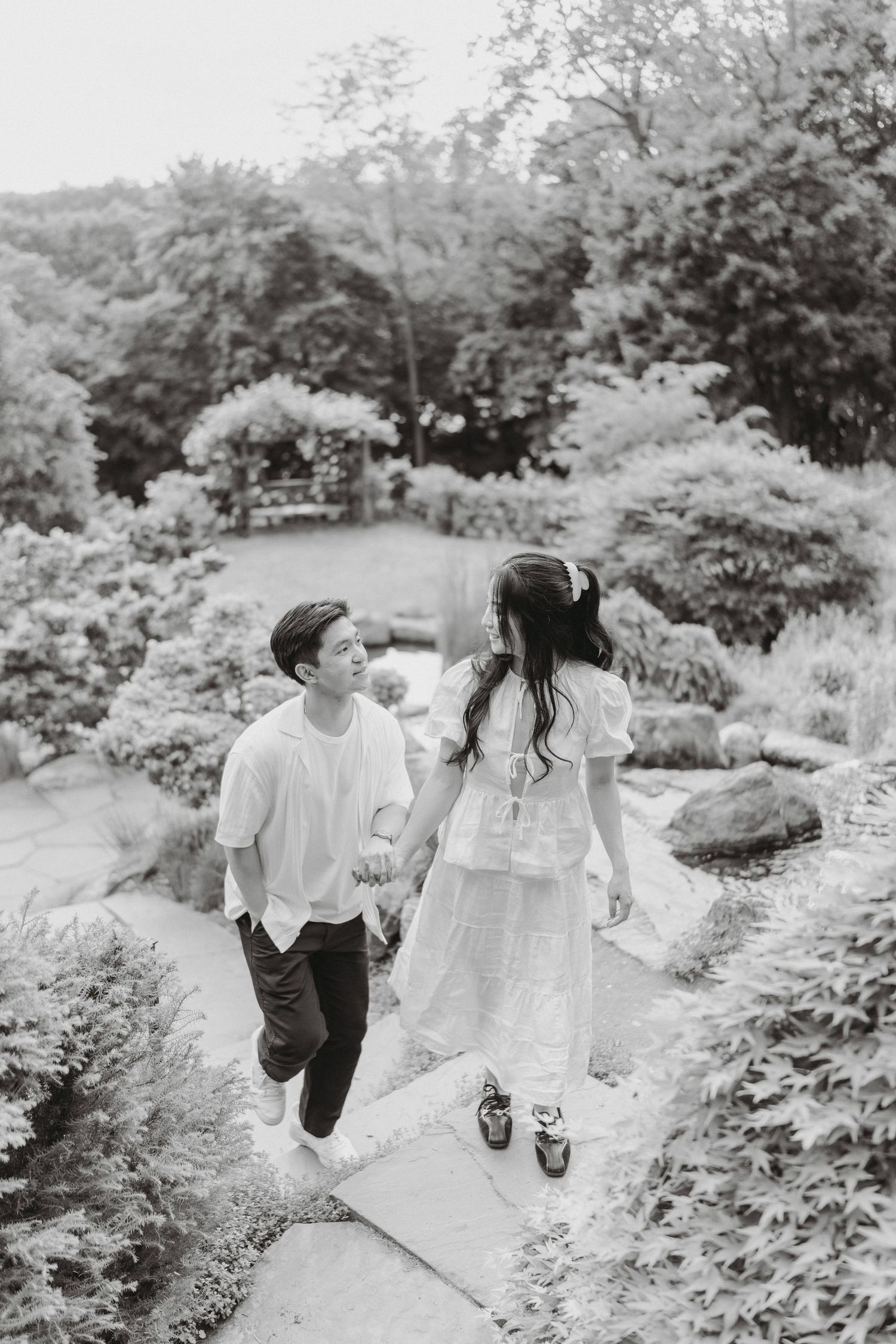 Black and white photo of a couple holding hands and walking up a stone garden path surrounded by lush shrubs and trees.