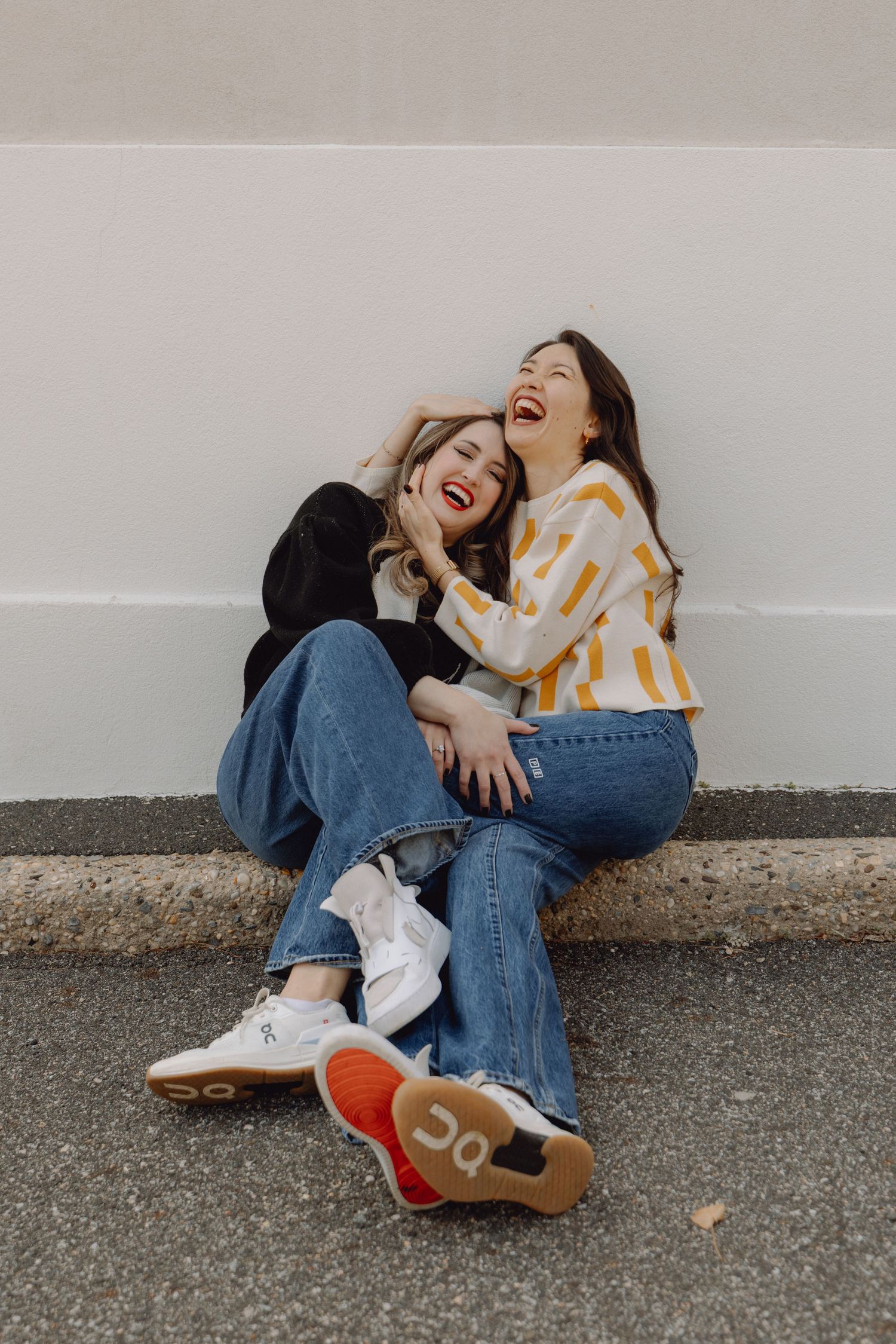 Two young women laughing and sitting together on a curb against a white wall, wearing jeans and casual tops.