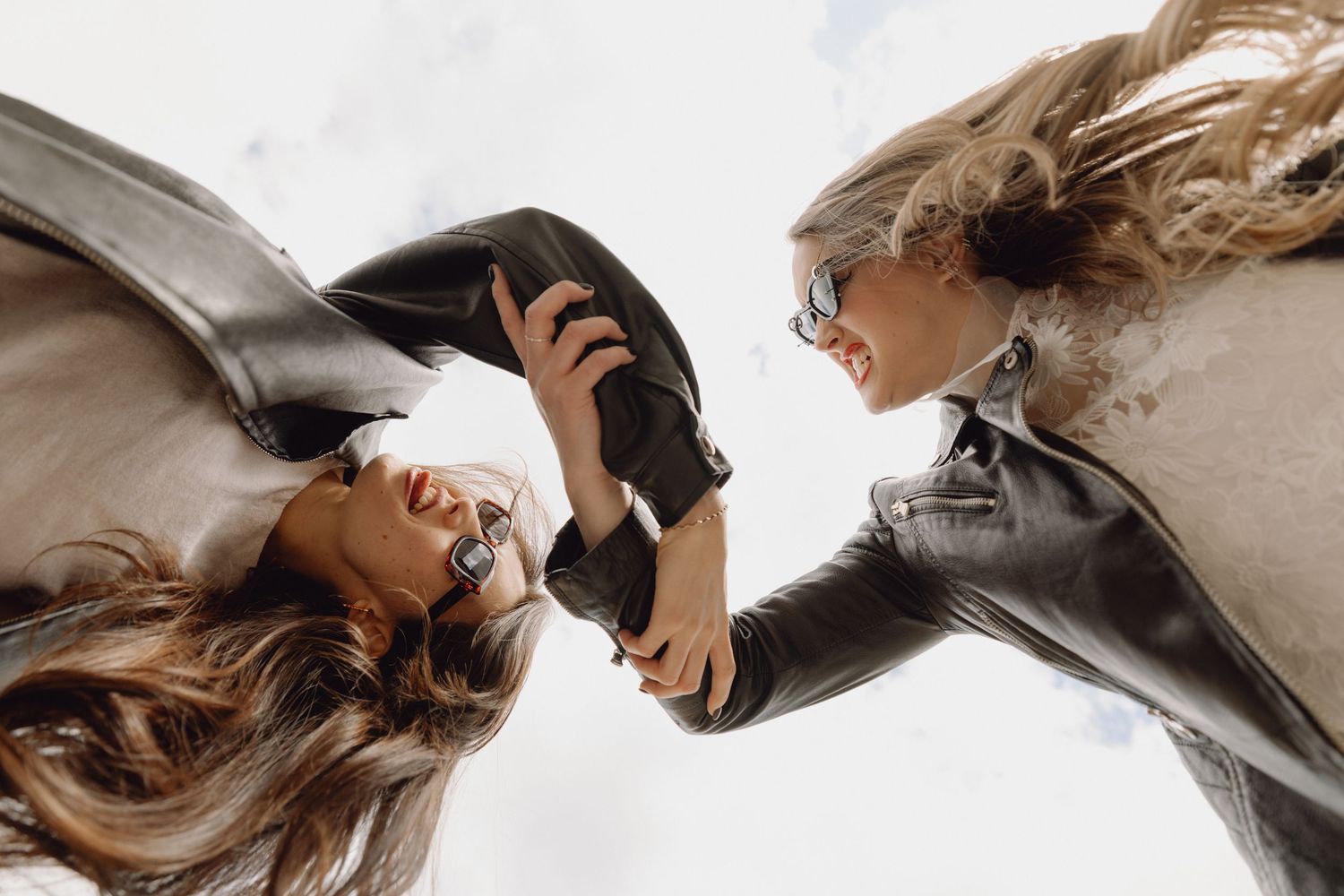 Two women in leather jackets and sunglasses face each other in a low-angle shot against a bright sky.